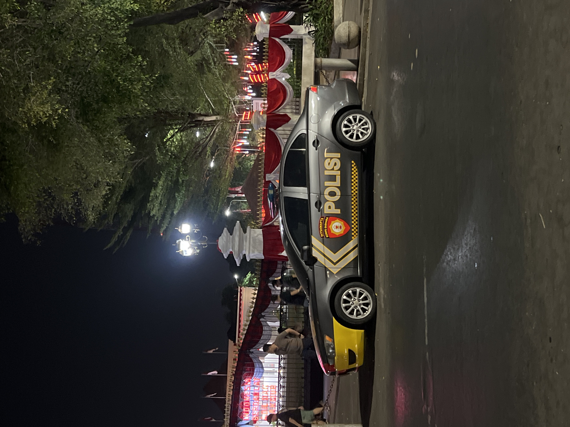 The image captures a night scene in front of the Gedung Agung Presidential Palace in Yogyakarta, Indonesia. A gray police car with "POLISI" and Indonesian national symbols visible on its side is parked on the street. The car has a yellow and black accent on its front bumper and a yellow stripe along its side. In the background, the Gedung Agung is decorated with red and white banners, Indonesian flags, and string lights. Several people are visible walking in the vicinity of the palace entrance, some appearing to be guards or officials. The environment suggests an event or a heightened security presence due to the decorations and the police car. The overall atmosphere is nocturnal, with artificial lighting illuminating the scene.