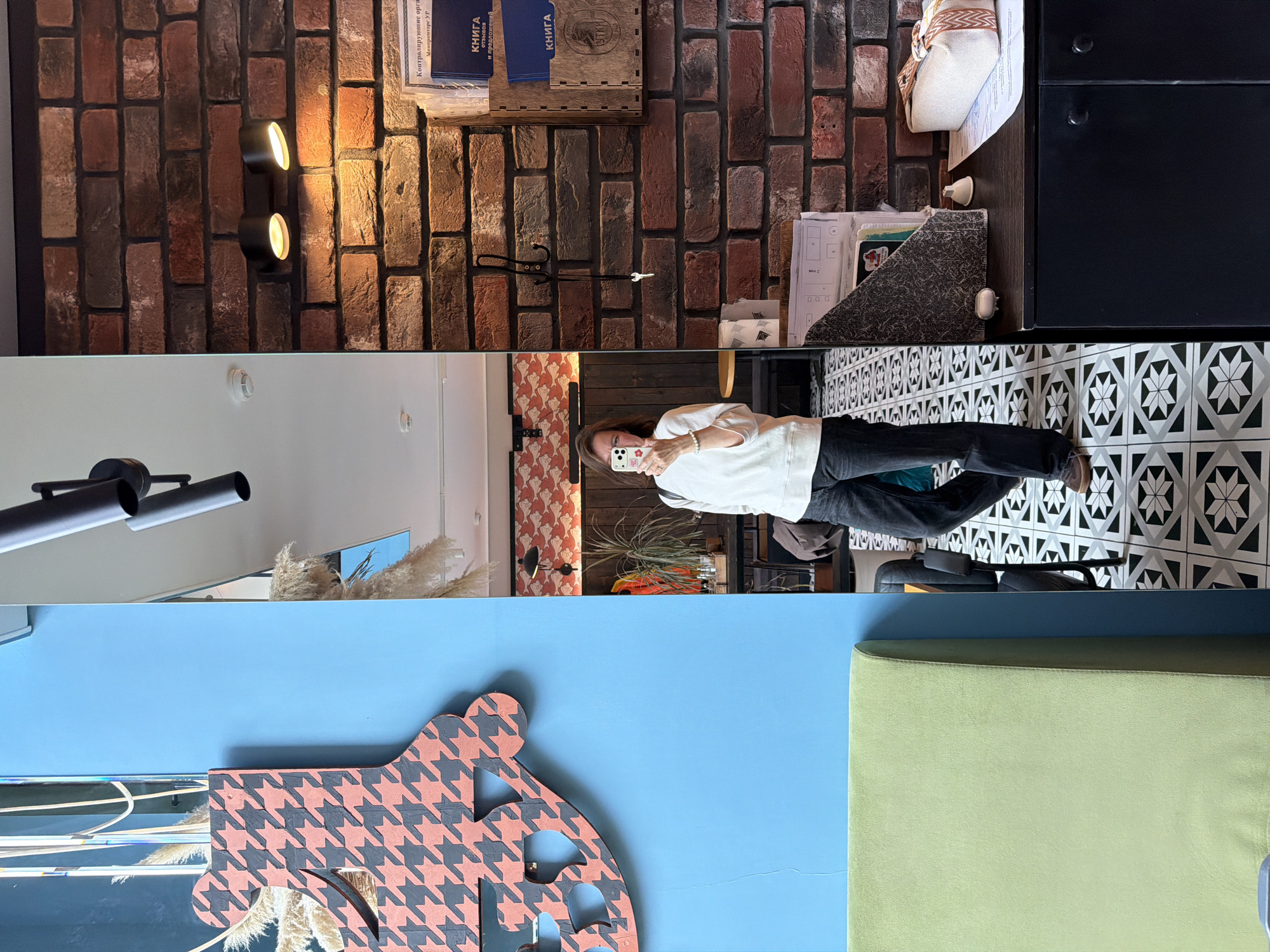 An interior shot depicts a woman taking a mirror selfie in a commercial establishment located in Izhevsk, Russia. The woman, with medium-length brown hair, wears a loose white long-sleeved top and dark jeans, and is holding a light-colored smartphone to capture her reflection. A light-colored bracelet is visible on her left wrist.
To her immediate left, a bright blue wall is visible, adorned with a large, abstract decorative element featuring a pinkish-orange and dark gray houndstooth pattern. Partially reflected in the mirror behind this decoration are dried ornamental grasses. A segment of a light green upholstered bench or seating unit is visible in the lower-left corner against the blue wall.
To her right, a prominent brick wall is featured, showcasing bricks in various shades of red, brown, and gray. Mounted on this wall are two black cylindrical spotlights emitting warm light downwards. Below these lights, a black three-pronged coat hook holds a small silver-colored key on its lowest peg. To the right of the hook, a wooden wall-mounted organizer contains several blue folders or books with white Cyrillic text, including "КНИГА" (Book) and "МИНПРОМТОРГ УР" (Ministry of Industry and Trade of Udmurt Republic), which identifies the location as being within the Udmurt Republic, with Izhevsk being its capital.
Below the organizer, a dark-colored counter or cabinet with two black handles supports various items: a textured dark brown magazine holder, a small white bell-shaped object, another small white irregular object, a stack of papers, and an elongated white fabric pouch with a striped orange and white handle.
The floor throughout the visible space features a geometric pattern of black, white, and gray tiles arranged in repeating star-like or compass rose motifs.
The mirror's reflection behind the woman reveals a patterned wallpaper with a repeating reddish-orange and light gray fish or cloud-like design, situated above dark wooden paneling. A tall green potted plant rests on a dark table, next to various small bottles. A black wall-mounted lamp is visible on the patterned wall in the reflection. The ceiling in the reflection is white, with a smoke detector or recessed light fixture visible.