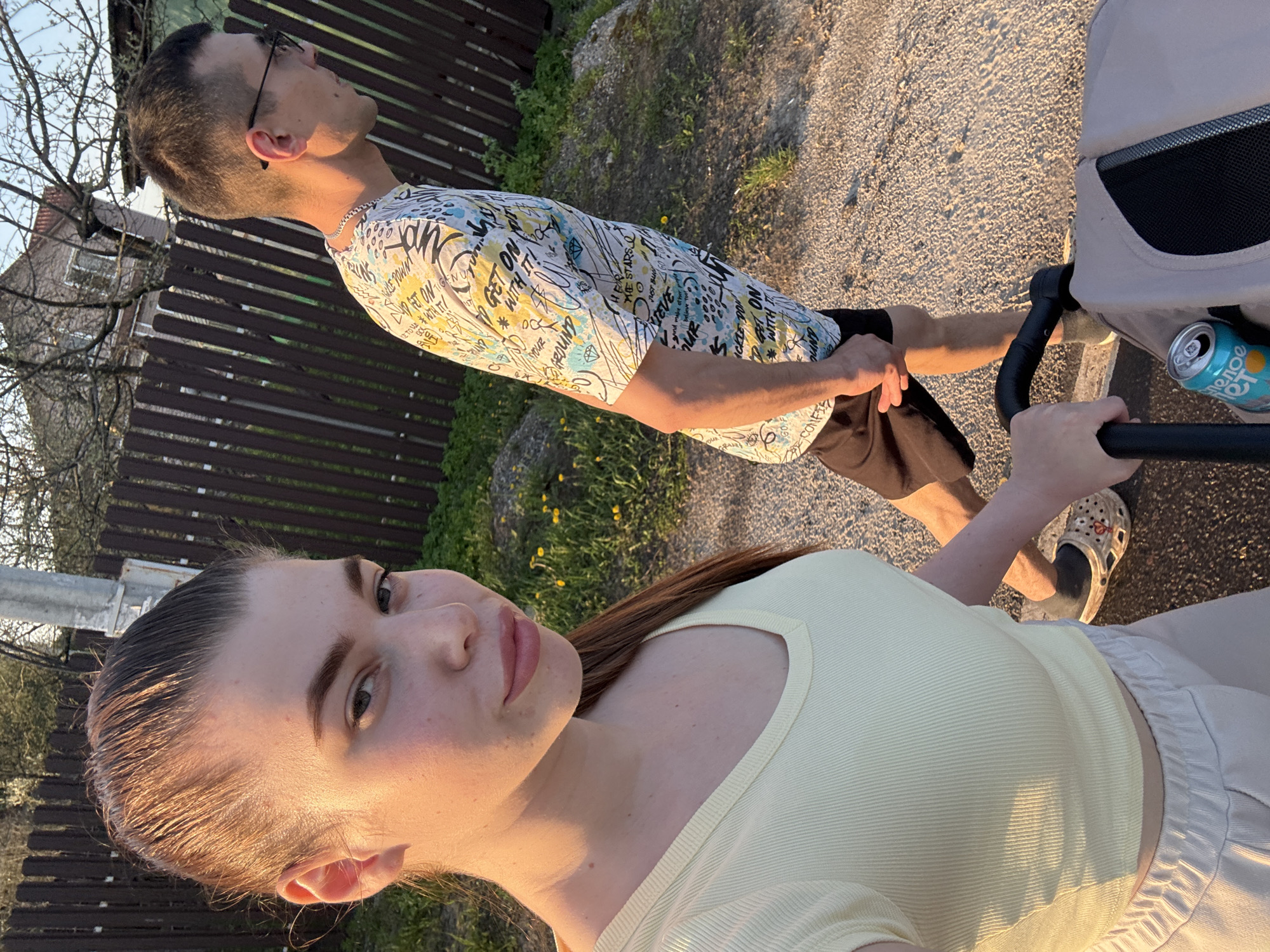 The image captures a family scene in Shevlyagino, Russia, featuring a couple strolling outdoors with a stroller. The woman is in the foreground, facing the camera, wearing a light yellow top. Behind her is a man in a patterned white t-shirt and dark shorts, with glasses, appearing to look upwards. The environment includes a gravel path, a wooden fence, and greenery. The stroller has a beverage can within reach. The lighting suggests it's late afternoon or early evening.