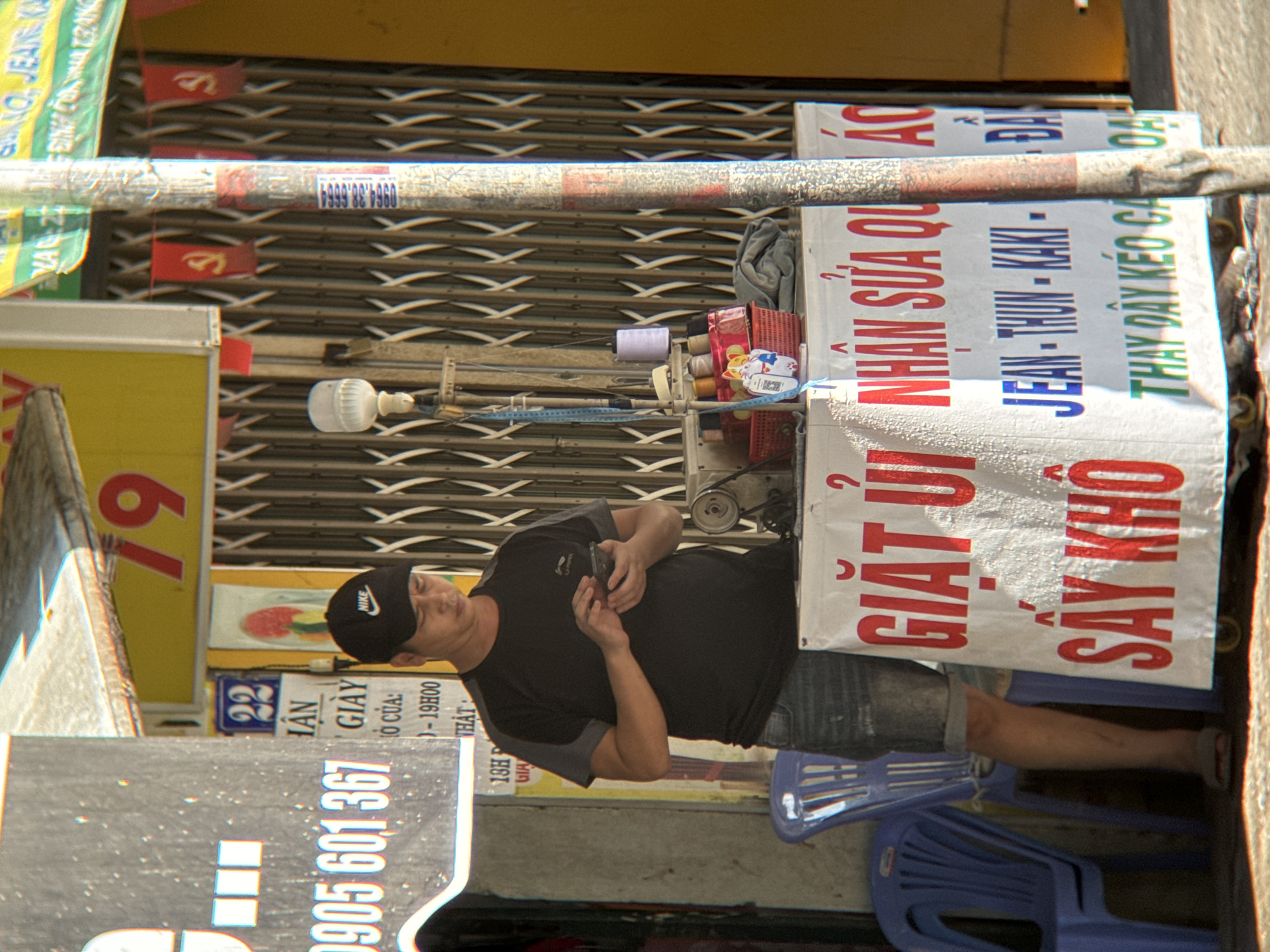 The image depicts a street scene in Nha Trang, Vietnam, likely in front of a shop. A man, wearing a black Nike cap and a dark t-shirt with jeans, stands with his phone in his hands. He is positioned in front of a white banner with red and blue lettering, which appears to be advertising services such as "GIẶT ỦI NHẬN SỬA QUẦN ÁO" (Laundry and Clothing Repair) and "SẤY KHÔ" (Dry Cleaning). Beneath this, further text lists types of clothing like "JEAN - THUN - KAKI" and "DÂY KÉO, DÂY..." (zippers, strings...). To the left of the man, a blue plastic chair is visible, and to his right, a sewing machine and various spools of thread are on display, reinforcing the notion of a repair or tailoring service. In the background, a metal grate-like security door is present. A utility pole with various posters and stickers is also prominently featured. A yellow awning with the number "79" in red is visible above. The overall impression is that of a small business operating on a city street.