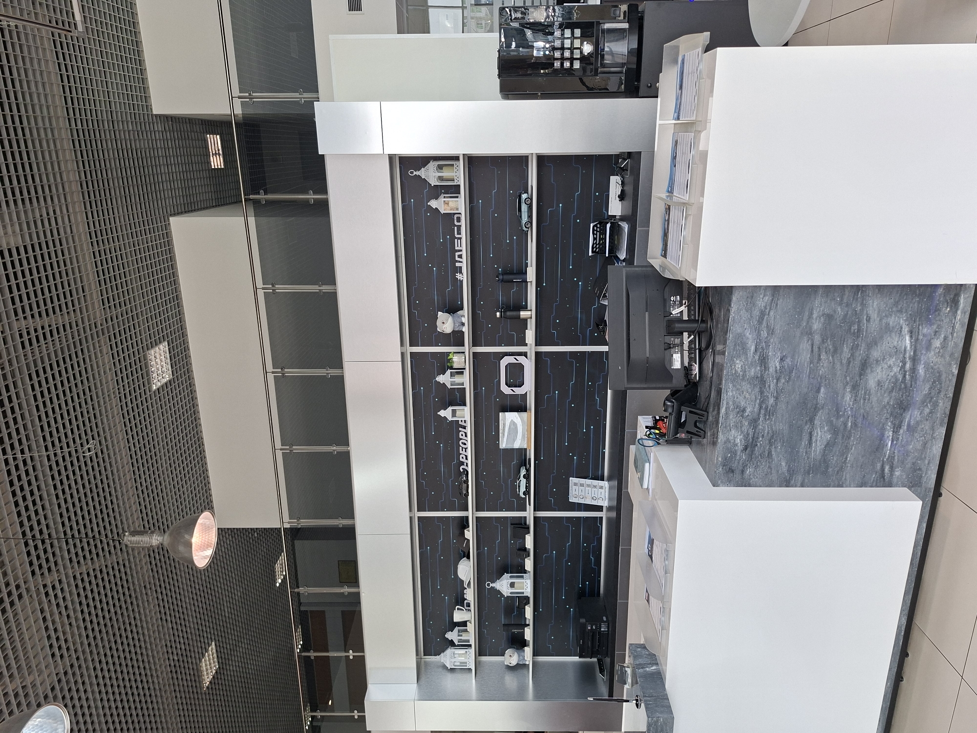 This is a photograph of the interior of a modern commercial space, possibly a dealership or a showroom, located in Izhevsk, Russia. The scene depicts a clean, well-lit environment with a prominent display shelf unit featuring a dark, patterned background with blue circuit-like lines. The shelves are organized with various decorative items and product displays, including white lanterns, a white mug, a white baseball cap, a plush toy dog, a black water bottle, and miniature car models. There is also a white sign with the text "#IAECO".
In the foreground, a white reception desk is visible, with a dark gray, marbled countertop. On the desk, a computer monitor, a printer, and some brochures are placed. To the right of the reception desk, a large, dark coffee machine is situated. The ceiling is a gray, tiled grid, with a prominent industrial-style pendant light fixture hanging down. The overall impression is one of a contemporary and professional setting. There are no people visible in the image.