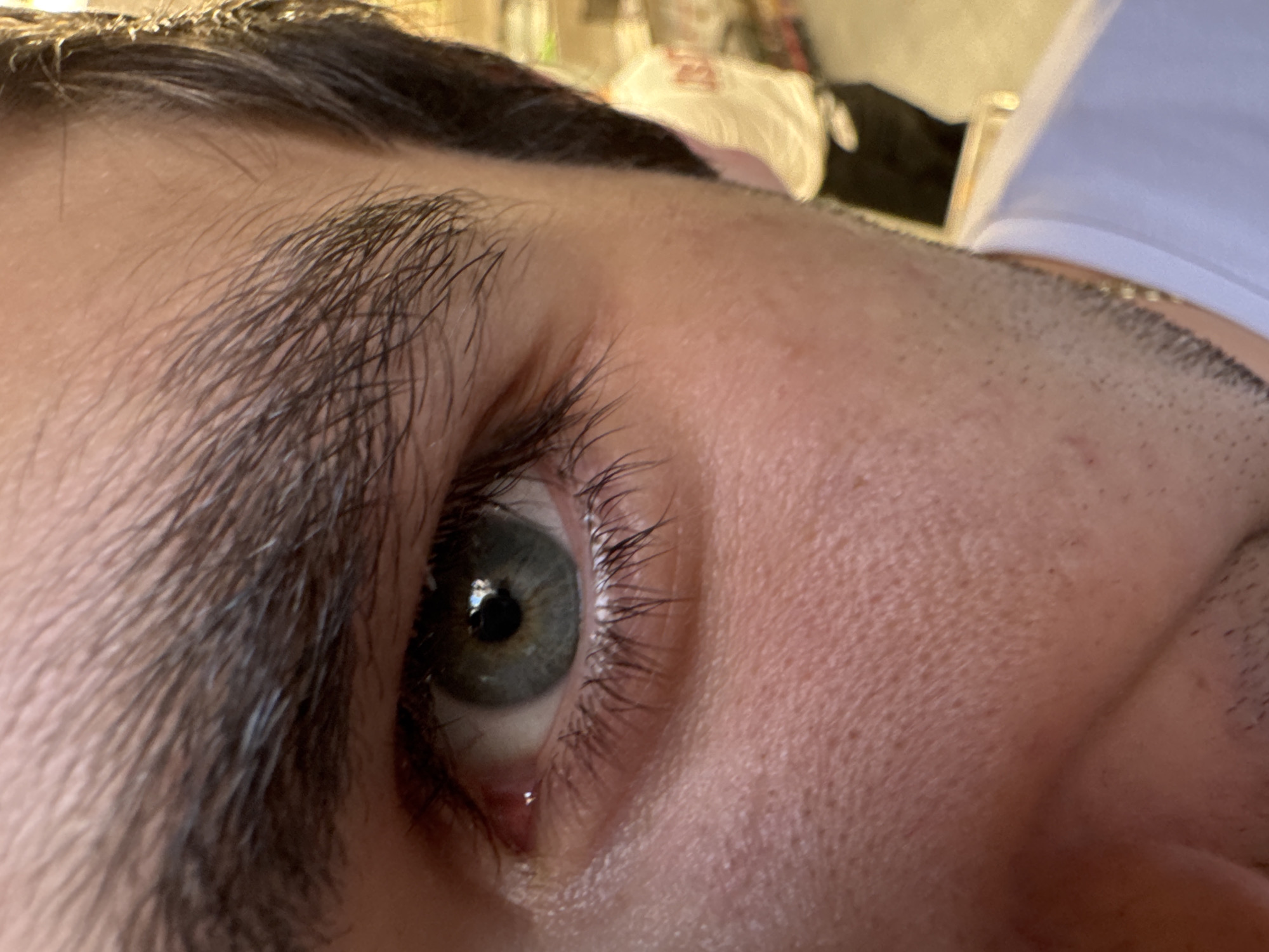 A close-up, angled shot focuses on a person's left eye and a portion of their face. The subject's eye features a hazel or greenish-grey iris with a dark pupil, reflecting what appears to be an indoor light source. Long, dark eyelashes frame the eye, and a prominent, thick dark eyebrow arches above it. The skin around the eye and on the forehead and cheek shows visible pores and fine hairs. There is a slight wetness in the lower tear duct of the eye.
The top and right sides of the image show parts of the person's head and neck area, including a hairline and a hint of stubble or very short hair near the ear/jawline. A thin gold chain is faintly visible on the neck. The background is heavily blurred but suggests an indoor setting with indistinct objects, possibly a white garment or bag. No specific activity is taking place beyond the close-up capture of the eye. There is no visible text in the image. The time of day appears to be daytime or well-lit indoors, and emotions are not clearly conveyed. The provided location context is Yerevan, Armenia, although no specific visual cues within the image confirm this.