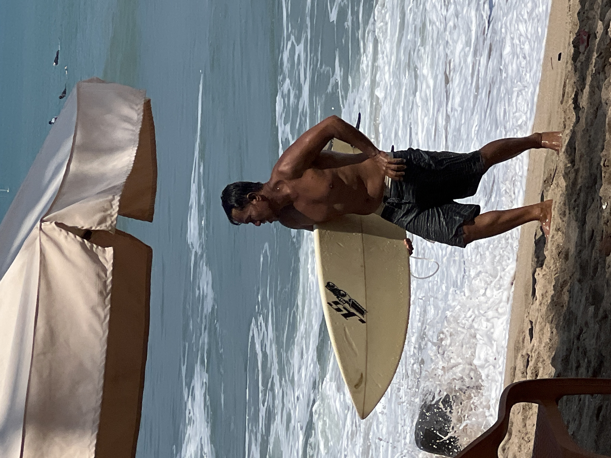 A shirtless man with dark hair and tanned skin walks on a sandy beach in Kuta, Indonesia, carrying a light-colored surfboard under his right arm. He is wearing dark, patterned board shorts. The man is facing towards the right of the frame, looking out at the ocean. The ocean is a vibrant blue with white foamy waves breaking on the shore. In the background, several other surfers can be seen in the water. The foreground shows a portion of a beach chair and the sand with shadows cast upon it. An umbrella provides shade over part of the scene.