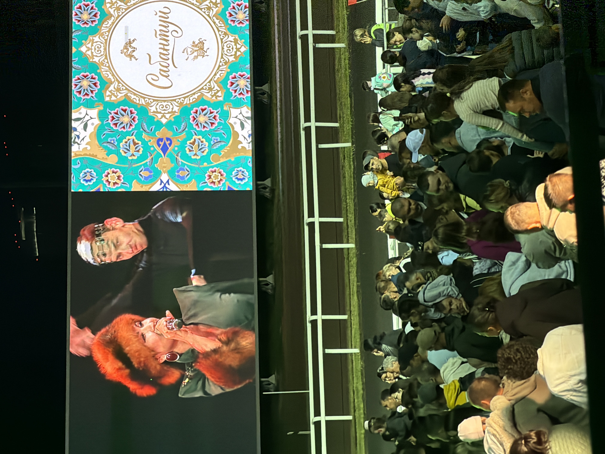 An outdoor event is captured at night, with a large screen displaying an image of a woman in a fur hat singing into a microphone on the left and a decorative pattern with text in a circular frame on the right. Below the screen, a white railing separates the event area from the audience, who are gathered in the foreground, facing the screen. The audience consists of a diverse group of people, including men, women, and children, some of whom are standing and others are sitting. The lighting suggests artificial illumination.