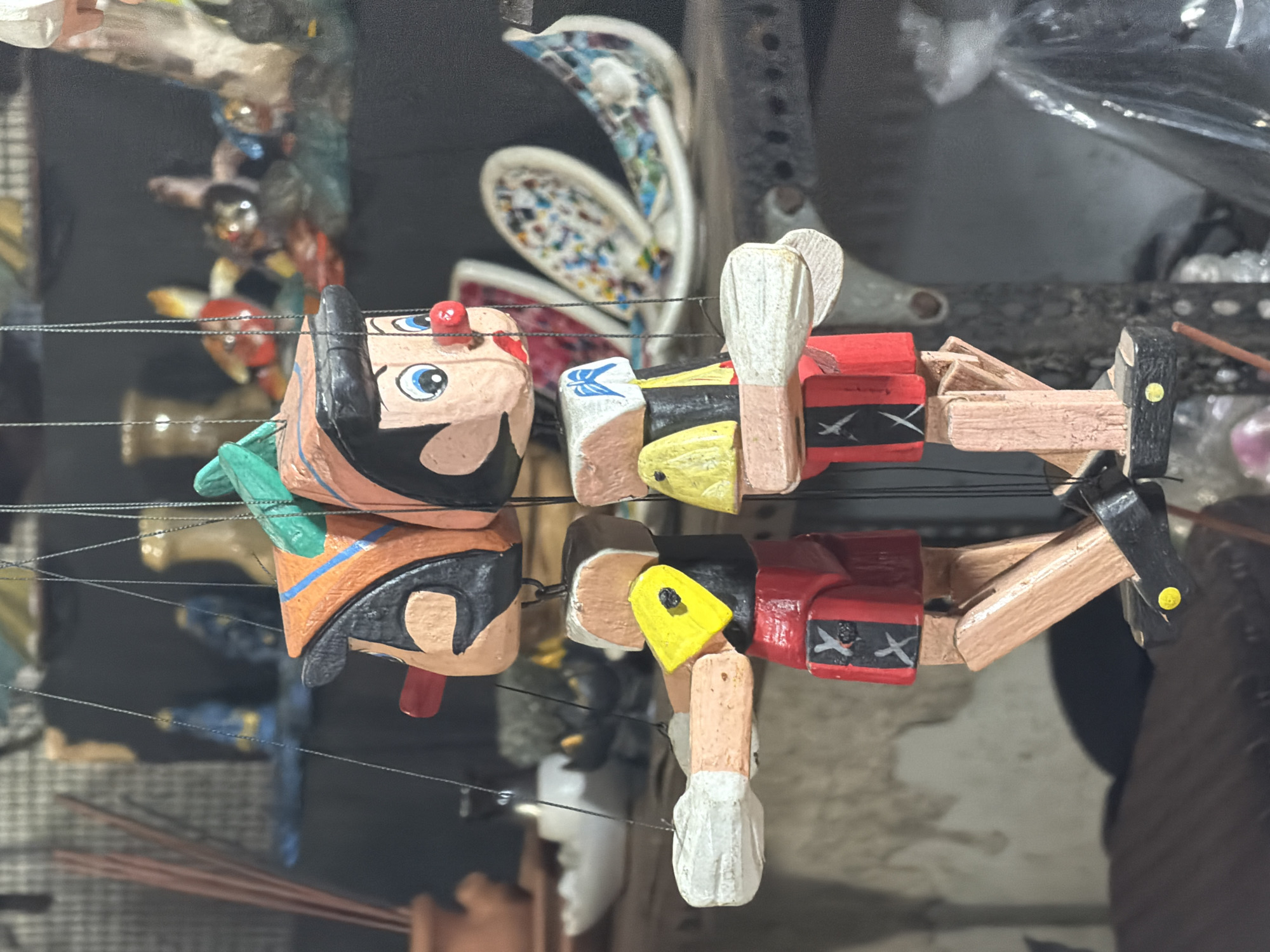 This is a close-up, vertically oriented image of a traditional Indonesian marionette, likely a Wayang Golek puppet. The puppet is made of carved and painted wood.
The subject is a single puppet, seemingly depicted in a shop or market stall. The puppet has a stylized human face with large blue eyes, black eyebrows, and a red nose. It wears a black wig and a headdress or hair ornamentation that is green on one side and orange with blue trim on the other. The puppet's body is segmented, made of cylindrical wooden pieces. Its torso is painted red with black and white decorative elements resembling crossed bands or ribbons. The arms are jointed, made of wood, and extend outwards, with one hand visible with a white glove or wrap. Thin black strings are visible, suggesting the puppet is suspended or ready to be manipulated.
In the background, other objects are blurred and out of focus, including what appear to be more puppets or craft items, as well as shelves and possibly plastic sheeting. The lighting appears to be artificial, typical of an indoor setting. There is no visible text in the image. The overall impression is of a handcrafted item displayed for sale. The location context of Kuta, Indonesia, suggests this puppet is part of the local artisanal crafts and cultural heritage.