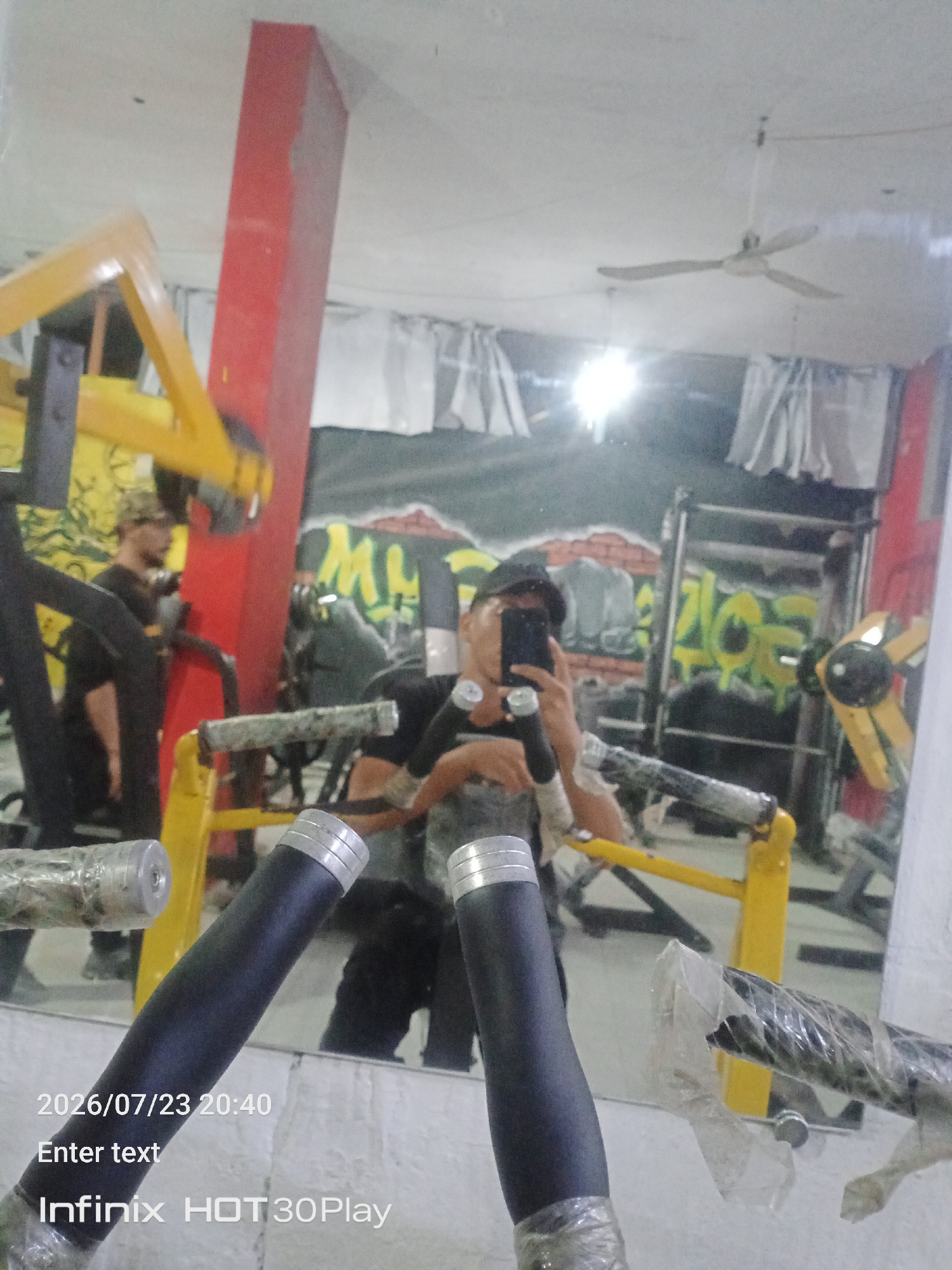 The image shows a man taking a selfie in a gym, reflected in a mirror. He is wearing a black t-shirt and a black baseball cap, and is holding a black smartphone in his right hand, angled to capture his reflection. The gym appears to be dimly lit and somewhat cluttered, with various weight training equipment visible in the background, including yellow and black machines and metal racks. Graffiti-like artwork is present on the walls behind the equipment. The floor is a light color, possibly concrete. The timestamp "2026/07/23 20:40" and "Infinix HOT30Play" are overlaid on the bottom left of the image, indicating the date, time, and phone model used to take the photo. A ceiling fan is visible in the upper portion of the mirror reflection. The overall impression is that of a casual snapshot taken by an individual in a personal fitness space, with no specific indication of its exact location within the city of Gaza, Palestinian Territories, beyond the fact that it is a gym.