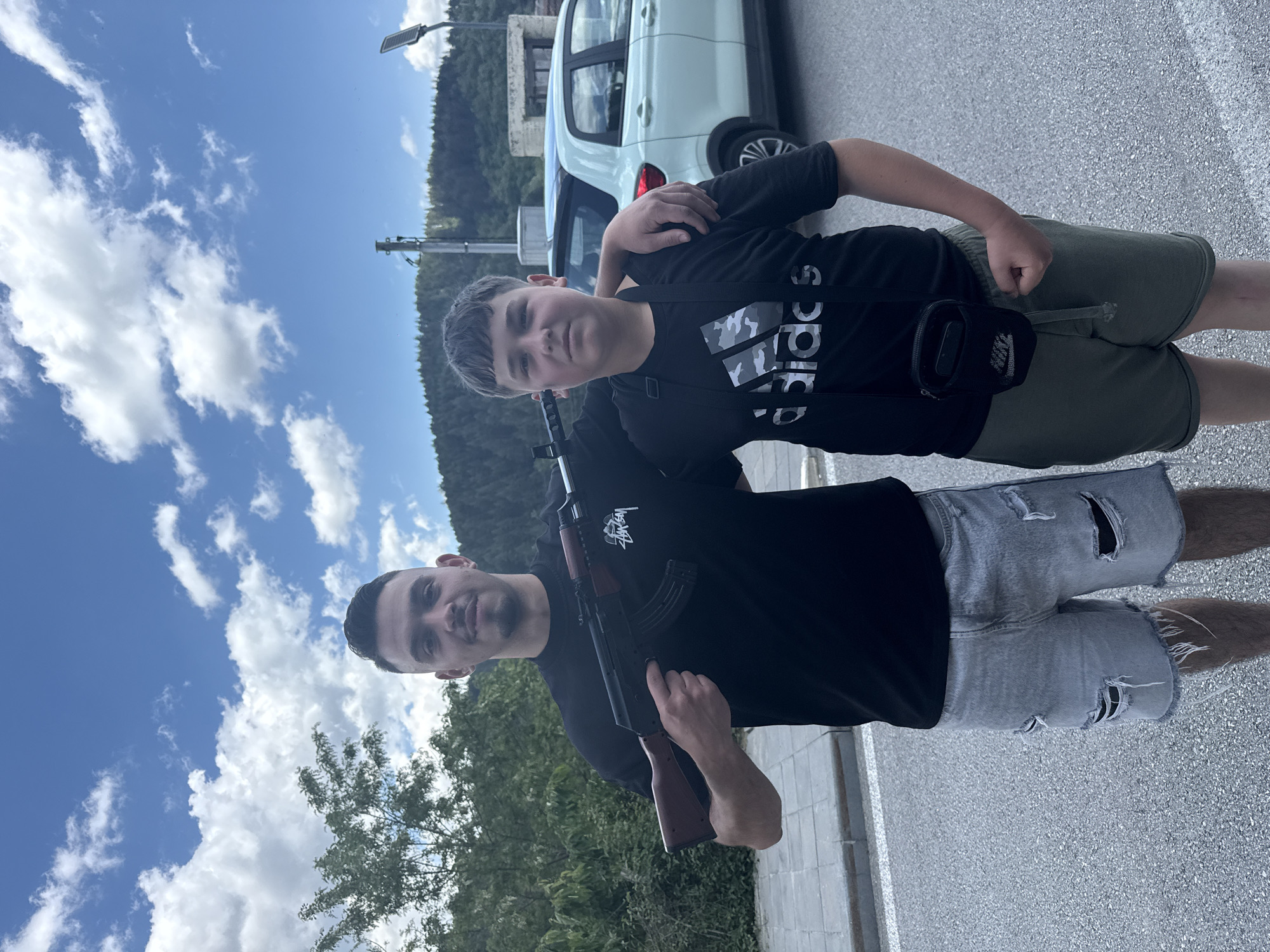 Two people stand outdoors, one holding an AK-47 style rifle. The person holding the rifle is an adult male wearing a black t-shirt and ripped denim shorts. A younger male stands next to him, wearing a black Adidas t-shirt and green shorts, with a black Nike side bag. The older male has his left hand on the shoulder of the younger male. In the background are trees, a light blue car, a building, and a sky with clouds. The ground is paved. The image was taken outdoors in Pripek, Bulgaria.