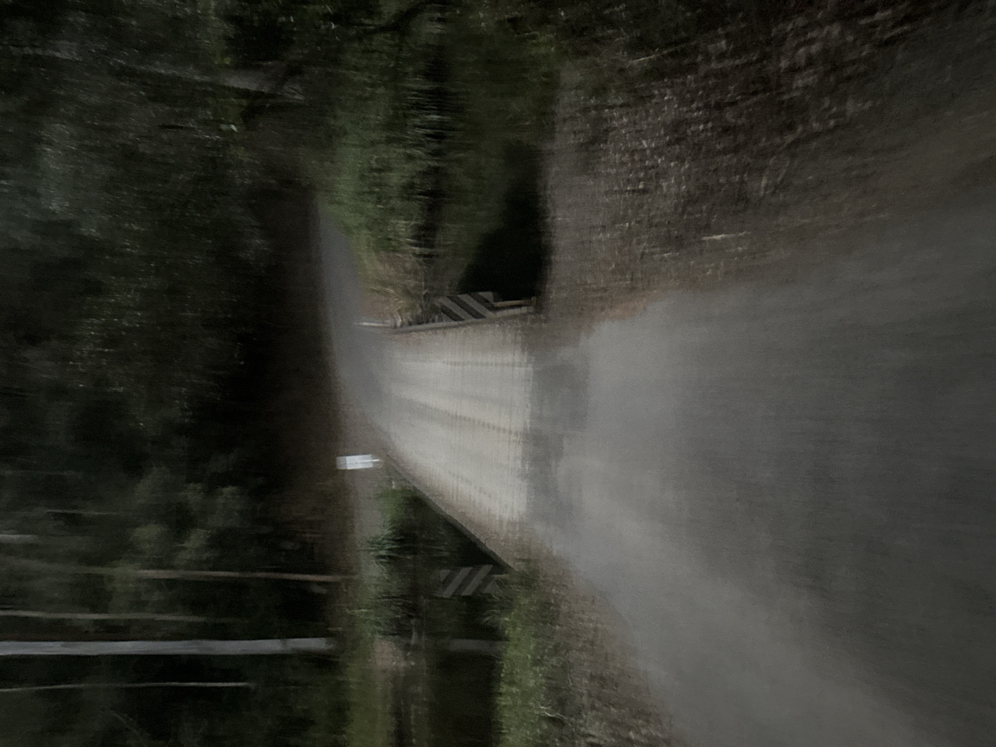The image shows a blurred, low-light view of a dirt road bridge crossing a body of water, likely a creek or slow-moving river, surrounded by trees and vegetation. The bridge appears to be made of dirt or gravel and has two distinct tire tracks visible on its surface. On either side of the bridge, there are wooden barriers with black and white diagonal stripes. The water is dark and reflects the surrounding trees, which are dense and form a dark backdrop to the scene. The overall impression is one of a rural or remote location, possibly a nature reserve or park, given the natural environment. There are no people or animals visible in the image. The location is identified as Thora, Australia, which suggests a setting in the hinterland region.