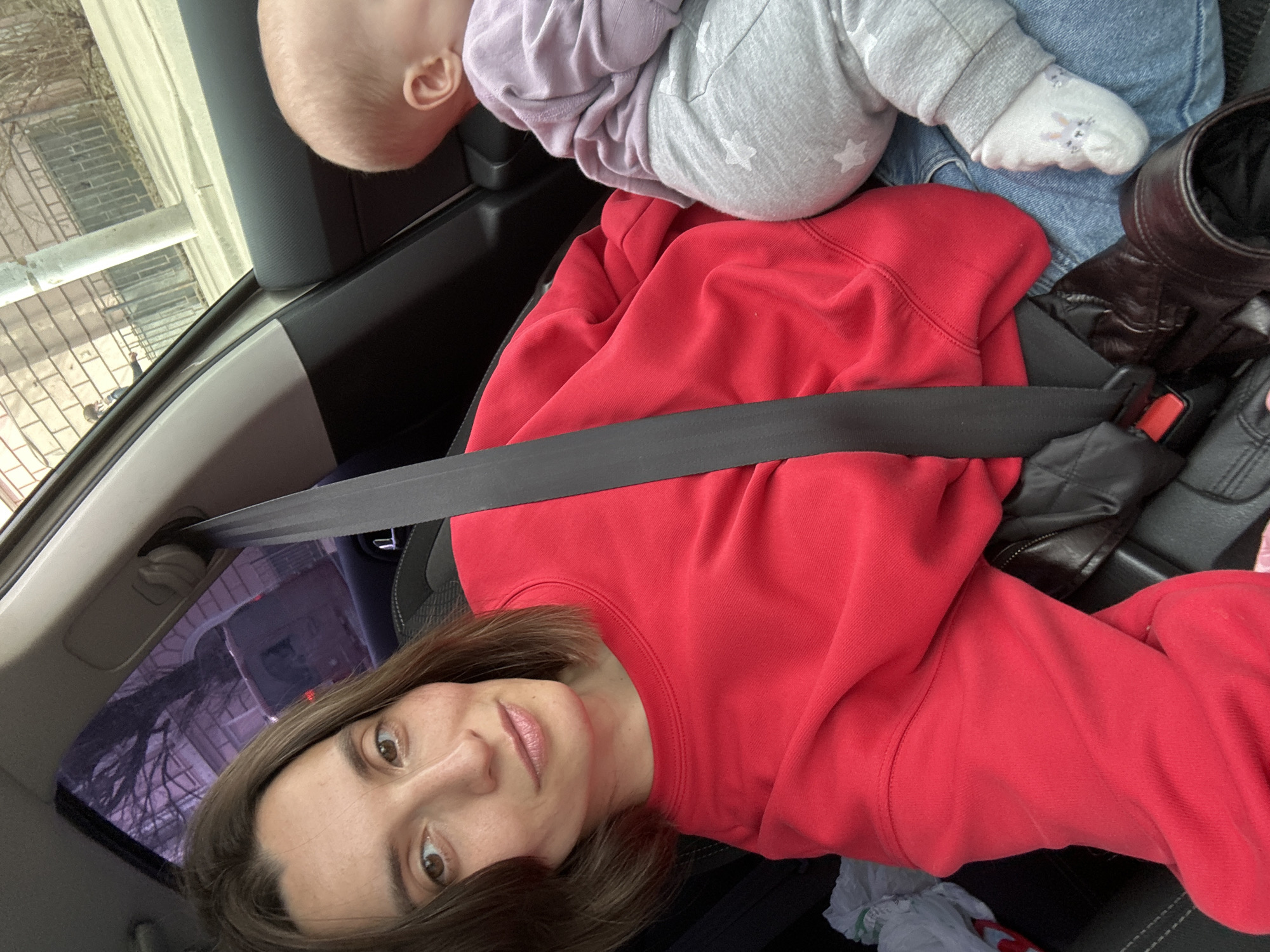 A woman is seated in a car, wearing a red sweatshirt and a seatbelt. A baby lies in the seat next to her, wearing a lilac top and gray pants with white stars, along with white socks. A black leather jacket lies on the seat. The car is traveling, and the scenery visible through the window is consistent with an urban environment, potentially Moscow, Russia, based on the architectural styles and overall urban density.
