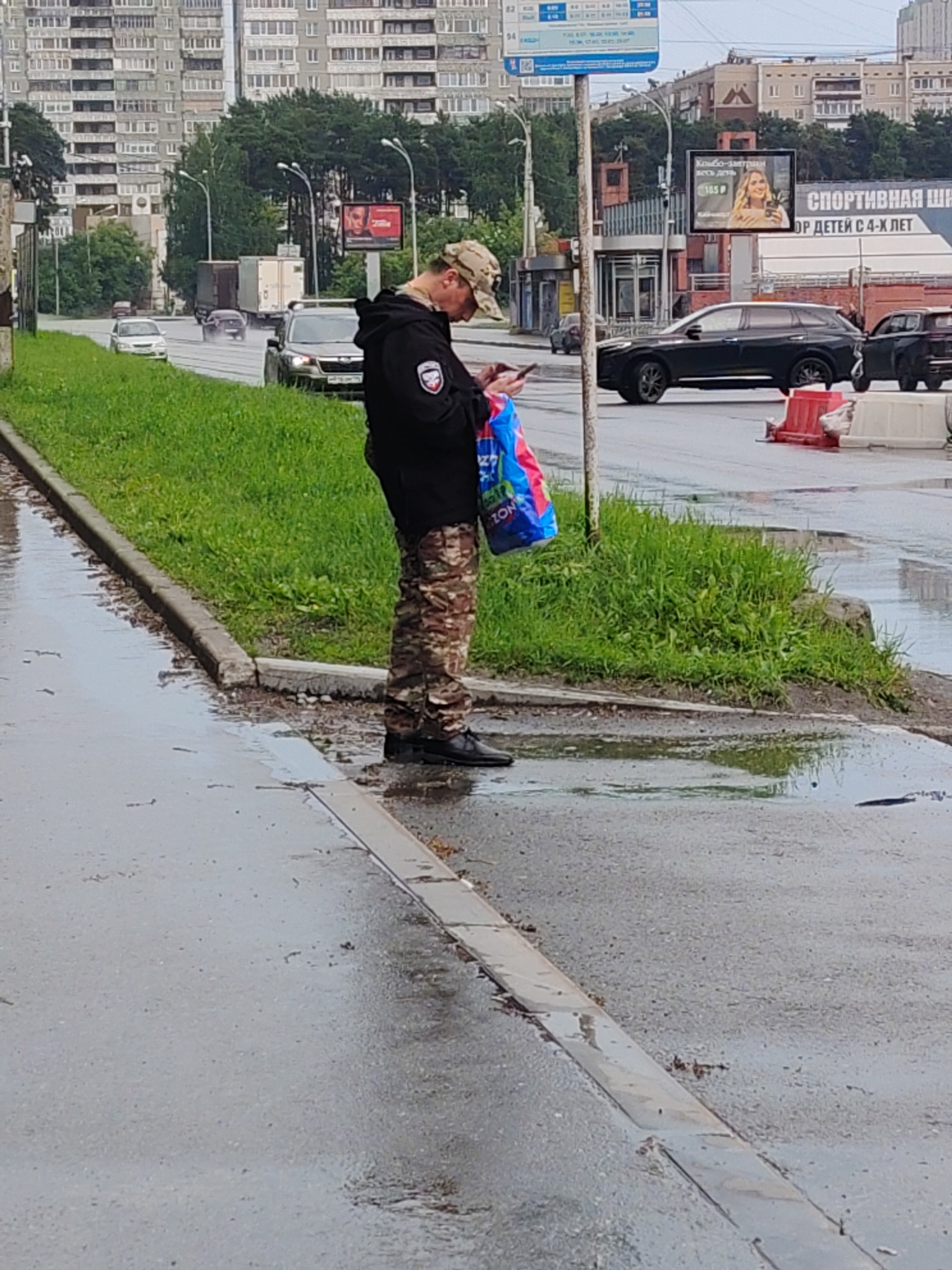 A person wearing camouflage pants and a dark jacket is standing on a wet sidewalk, looking at their phone. They are holding a blue and red shopping bag. The background shows a street with cars, a billboard with an advertisement, and tall apartment buildings. The scene appears to be in Yekaterinburg, Russia, based on the Cyrillic text on the billboard and the architectural style of the buildings. The weather is overcast and the ground is wet, suggesting it may have recently rained.