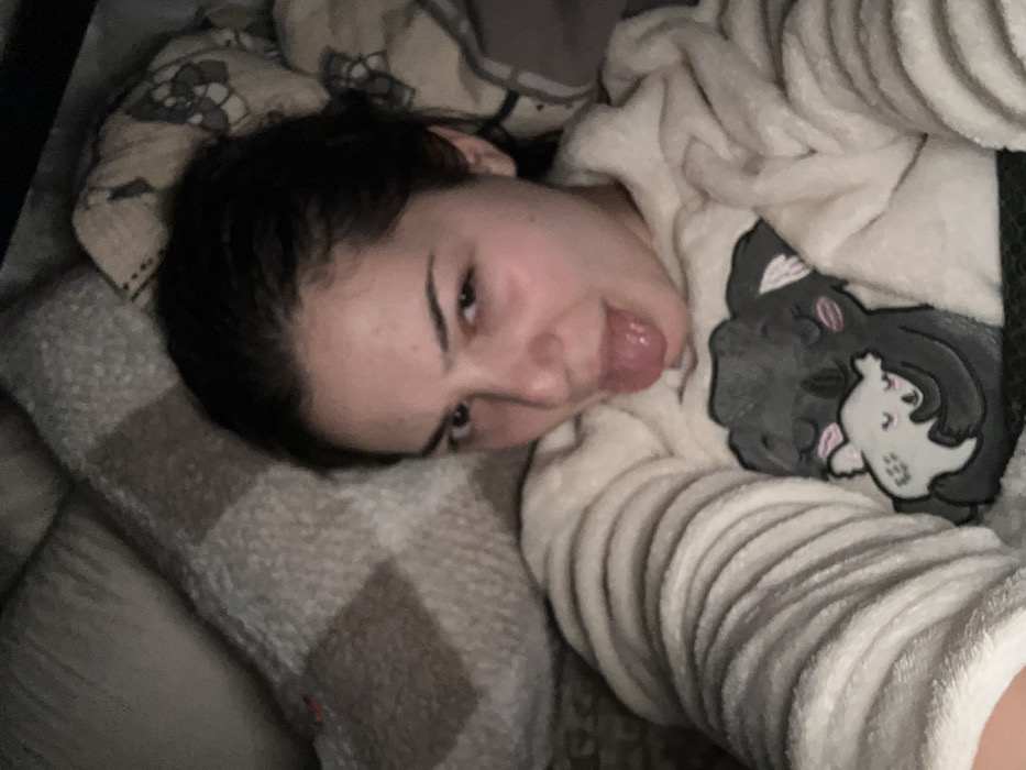 A young woman reclines in bed, her head resting on a plaid pillow. She wears a light-colored, fuzzy pajama top featuring a cartoon cat design. Her dark hair is pulled back, and she looks at the camera with a playful expression, sticking her tongue out. The scene appears to be indoors, softly lit and comfortable. The background includes a patterned blanket or bedding.