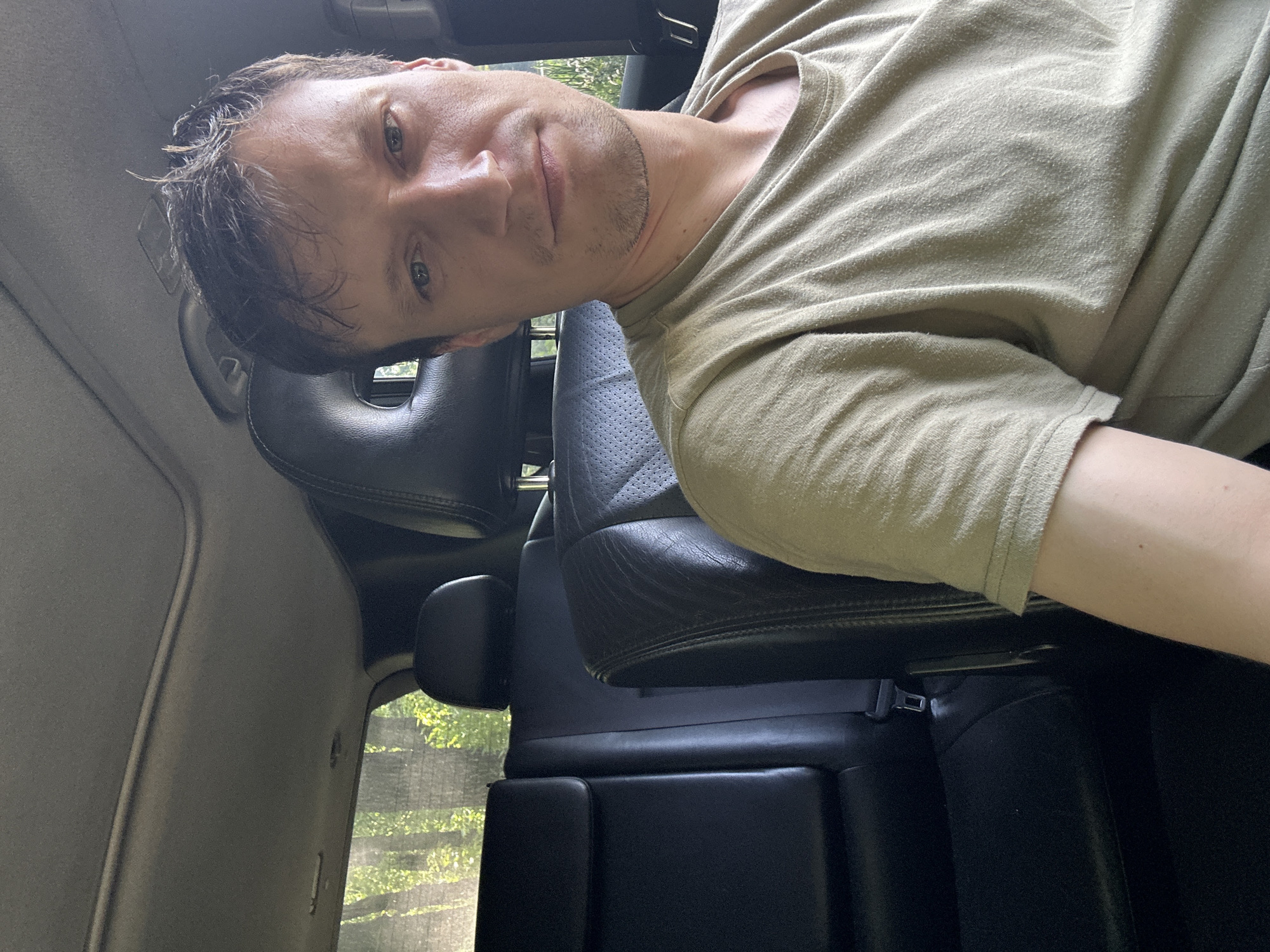The image is a selfie taken from the passenger seat of a car, looking towards the driver. The individual in the photo is a male with light skin, short brown hair, and a light stubble beard. He is wearing a light green t-shirt. Behind him, the interior of the car is visible, including black leather seats, headrests with circular cutouts, and a sunroof. Through the rear window, greenery and trees can be seen, suggesting an outdoor, possibly rural setting. The overall lighting indicates it is daytime. The location is identified as Makiv, Ukraine.