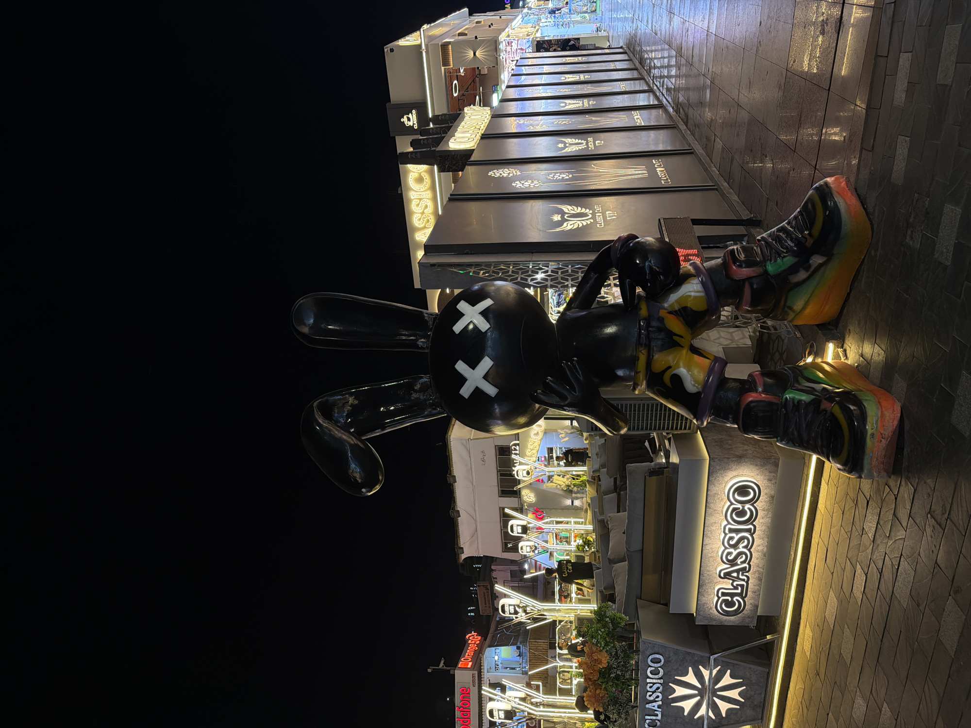 This is a night-time, eye-level, full shot of a large, black, cartoonish bunny statue with "X" eyes, located outside in Sharm El Sheikh, Egypt. The statue stands on a cobblestone walkway, which is wet, indicating recent rain. To the left of the statue, there is a seating area with plush couches and tables, illuminated by decorative white lights. Behind the seating area is a building with multiple storefronts, including one with a "CLASSICO CAFE" sign. Further to the left, a red "Vodafone" sign is visible. The statue is wearing colorful, patterned shorts and oversized, multi-colored sneakers. To the right of the statue, a metal fence with illuminated floral designs runs along the walkway, with more storefronts visible beyond it. The overall scene suggests an outdoor dining or entertainment area.