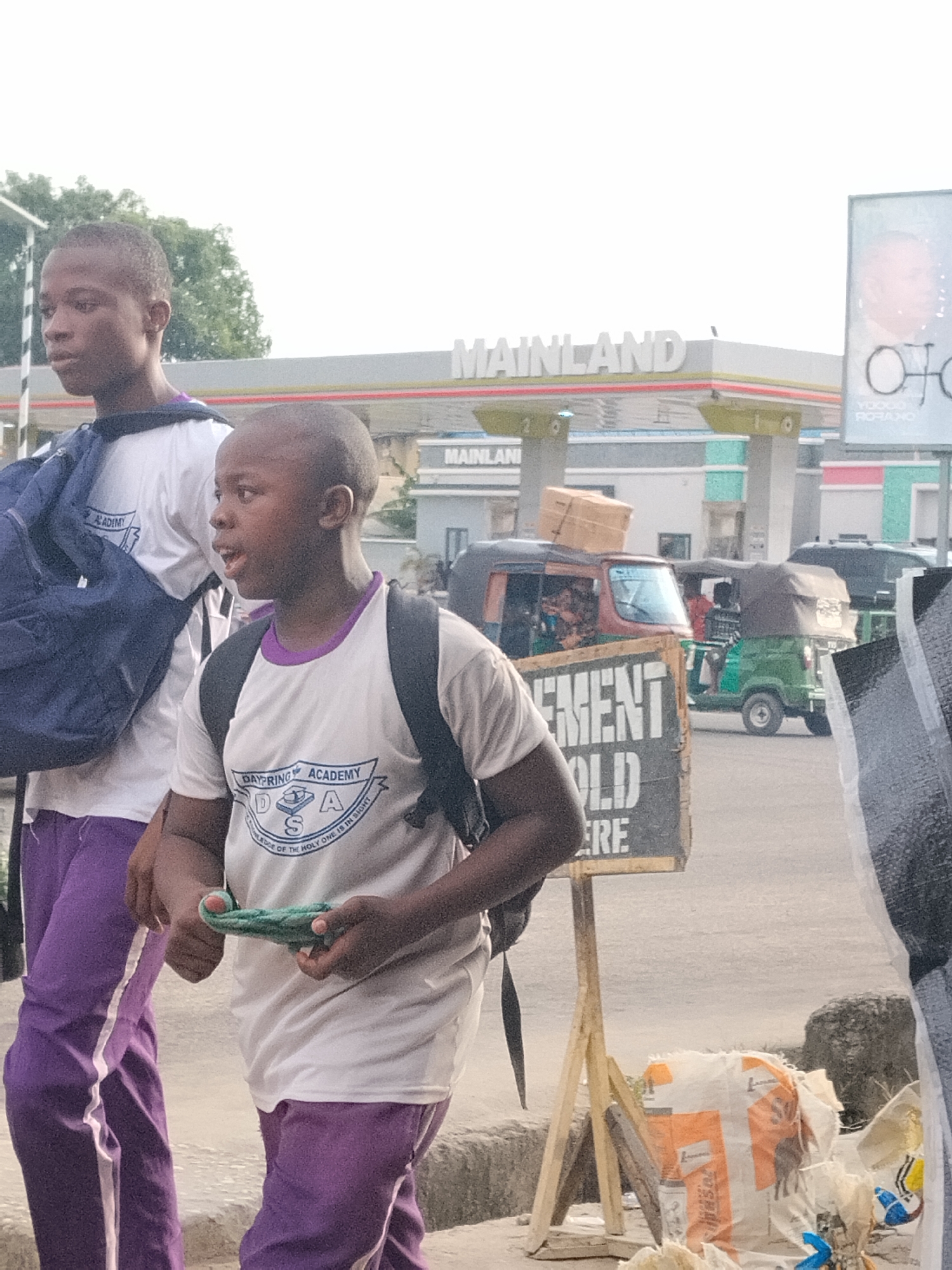 Two young boys, likely schoolchildren, walk along a street in Aba, Nigeria, during bright daylight. Both are dressed in white t-shirts with purple collars and trim, purple trousers with white side stripes, and black backpacks. The t-shirts bear the logo "DAYPRING ACADEMY" and the motto "KNOWLEDGE OF THE HOLY ONE IS IN SIGHT". The boy in the foreground holds a green, woven object. His companion walks beside him, also carrying a backpack that shows the word "ACADEMY".
The setting appears to be a bustling urban environment. In the background, a "MAINLAND" gas station is prominently visible with its canopy and pumps. Further right, a large billboard displays a person's face and text including "YODOO RORANO". On the road, several auto-rickshaws, commonly known as tuk-tuks, are present, one orange-brown carrying cargo on its roof, and another green one parked further back.
In the foreground, partially obscuring the view, is a wooden A-frame sign with fragmented text "EMENT", "OLD", and "ERE", suggesting it advertises cement sales. Next to it, on the ground, are several bags, some white and orange, appearing to contain building materials like cement. The overall scene captures daily life on a street, likely at a time when children are commuting to or from school.
