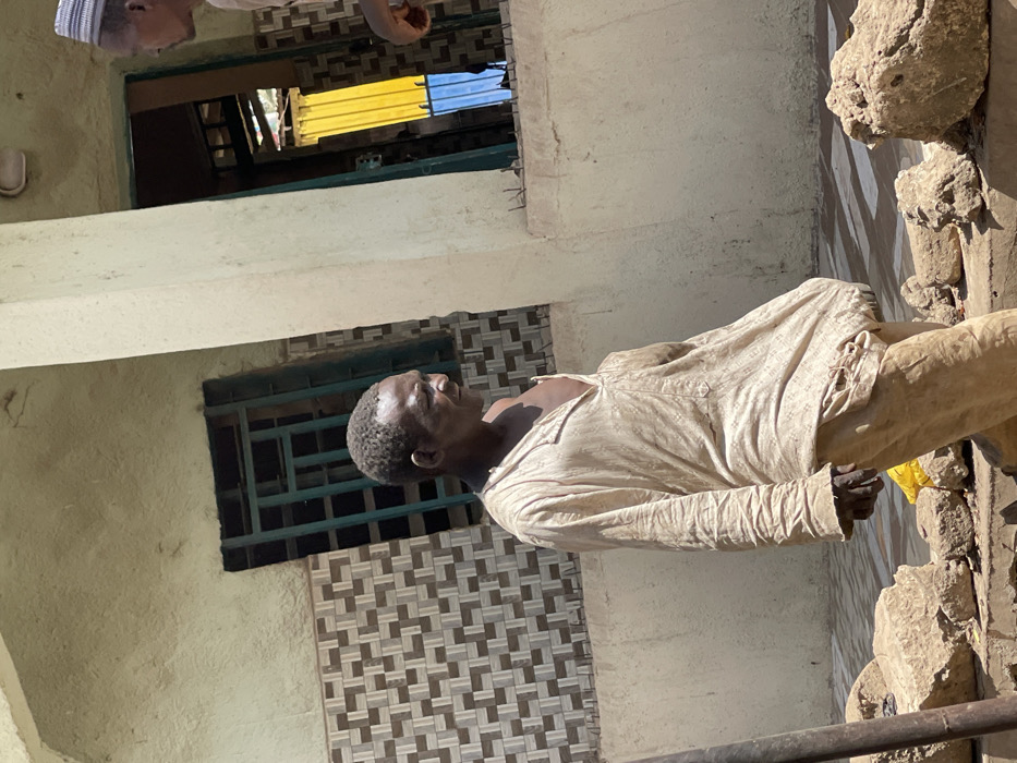 The image depicts an outdoor scene in Zaria, Nigeria, featuring two adult males against a building facade. The primary subject, a male with dark skin and short, dark hair, is positioned slightly right of center, facing right in profile. He wears a light-colored, long-sleeved tunic, open at the neck, revealing his chest, and light-colored trousers. His expression is neutral. To the far right, partially visible, is a second male wearing a light-colored cap and holding a reddish-brown object in his hand.
The background consists of a light-colored building wall with a lower section featuring patterned brown and off-white geometric tiles. Two window or door openings are present: one on the left with dark green metal grilles, and another on the right revealing a vibrant yellow structure and a blue surface deeper inside. A light-colored structural pillar separates the two individuals. The foreground is an unpaved, uneven surface with several large, irregularly shaped earth clods or rocks and a partially visible yellow object. The scene is brightly illuminated by direct sunlight, casting distinct shadows.