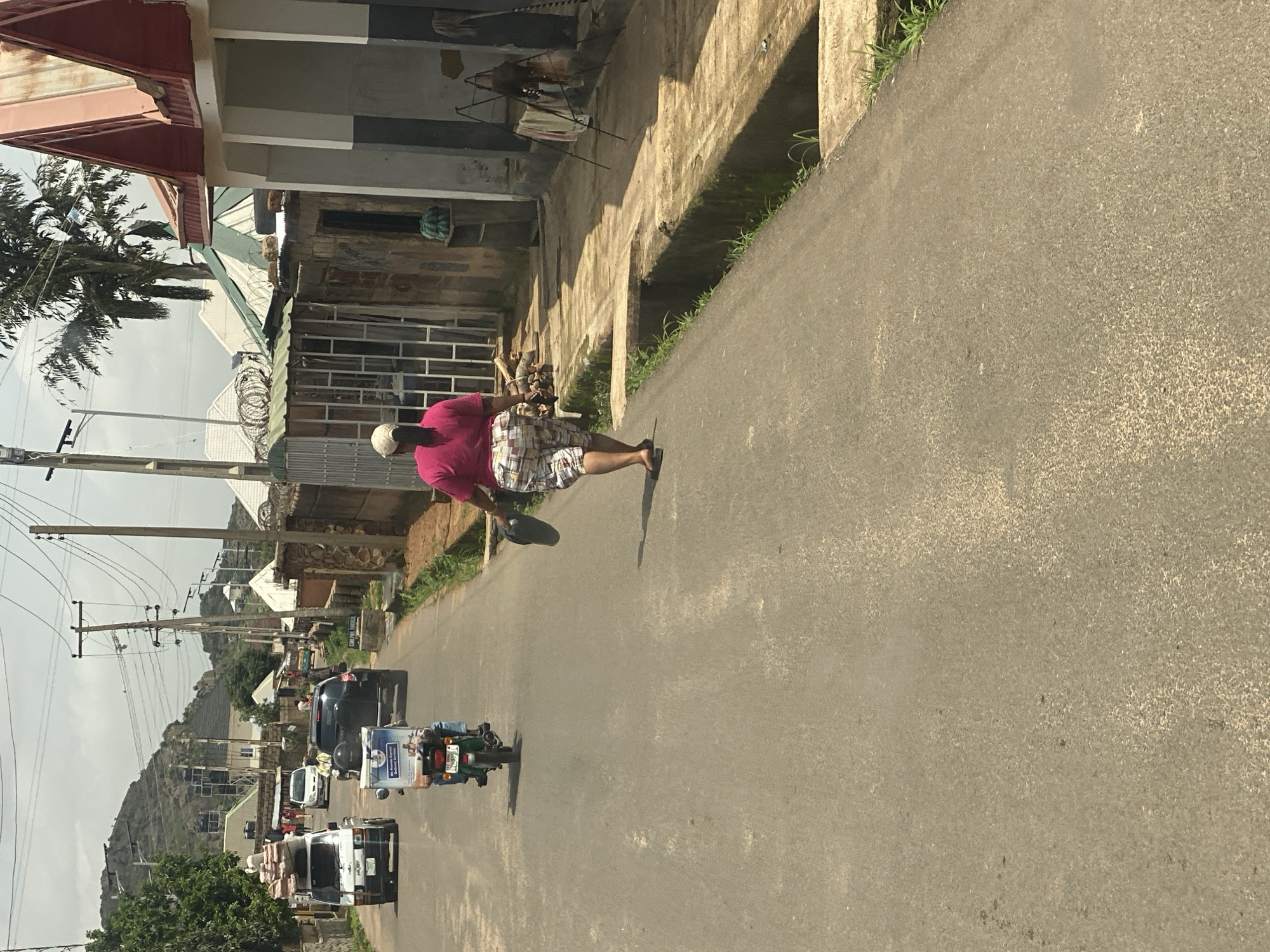 The image depicts a street scene in Barakin Akawo, Nigeria. The primary subject is a person walking on the right side of a paved road. The person is wearing a pink shirt and plaid shorts, with a white cap or head covering. They appear to be walking away from the camera, and their shadow is cast on the road.
The setting is a street lined with buildings on the left. Some of these buildings appear to be residential or commercial, with various architectural styles visible, including corrugated metal roofs and brickwork. There is a visible drainage ditch running alongside the road in front of some of the buildings. Power lines and utility poles are also present along the street.
Further down the road, several vehicles are visible, including a black car, a white van carrying goods, and a motorcycle with a rider and a passenger. The motorcycle has some text on its rear panel, but it is not clearly legible. The road itself is asphalt and appears to be in fair condition.
The time of day appears to be daytime, as indicated by the shadows and the lighting. The weather seems to be overcast or hazy, as the sunlight is diffused. The overall atmosphere is that of a typical day in a Nigerian town, with activity on the street and evidence of everyday life. There are no explicit indicators of a specific event, but rather a snapshot of a normal day.