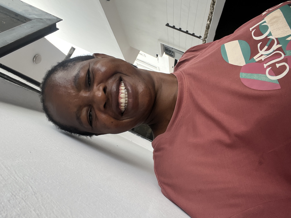 A front-facing, waist-up portrait captures an adult female with dark skin and short, dark hair, smiling broadly with visible teeth, leaning against a white textured wall. She is wearing a salmon-colored short-sleeved t-shirt featuring a multicolored "GUCCI" logo on the chest, designed with geometric shapes and stripes in pink, teal, white, and light brown hues. The background to her right reveals an outdoor setting under bright daylight. Visible elements include a dark, angled overhead architectural beam or roofline, a distant white building, and a dark perimeter fence equipped with security wires or spikes. The scene is located in Lekki, Nigeria.