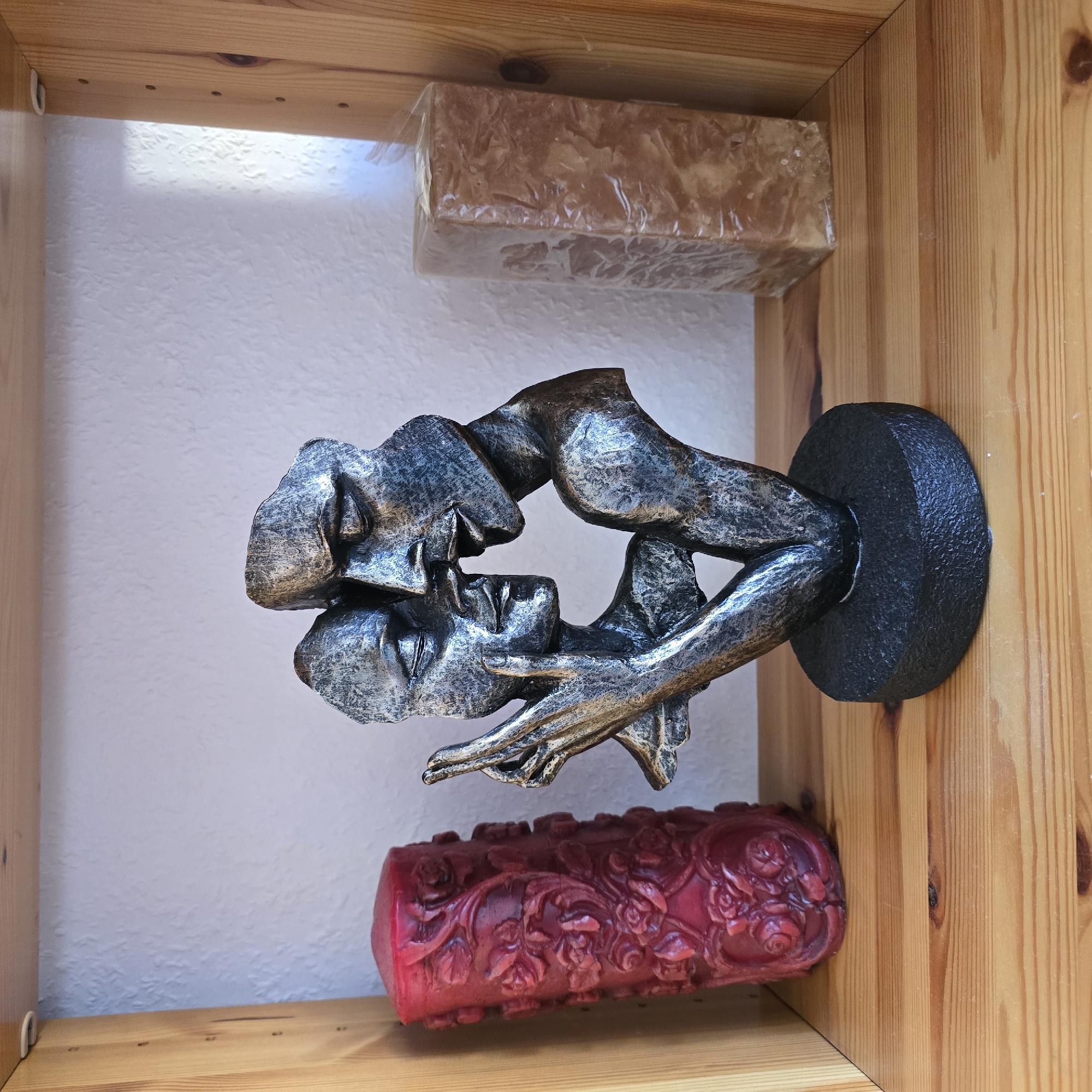 The image shows a wooden shelf in what appears to be a home in Zeithain, Germany. On the shelf are three decorative items. On the left is a tall, red cylindrical candle with an ornate, floral pattern carved into its surface. In the center is a metallic sculpture depicting two abstract faces in an embrace, with one face touching the other's cheek. The sculpture is mounted on a round, black base. On the right is a rectangular, translucent object, possibly another candle, wrapped in clear plastic. The background is a textured white wall, and the shelf itself is made of light-colored wood.
