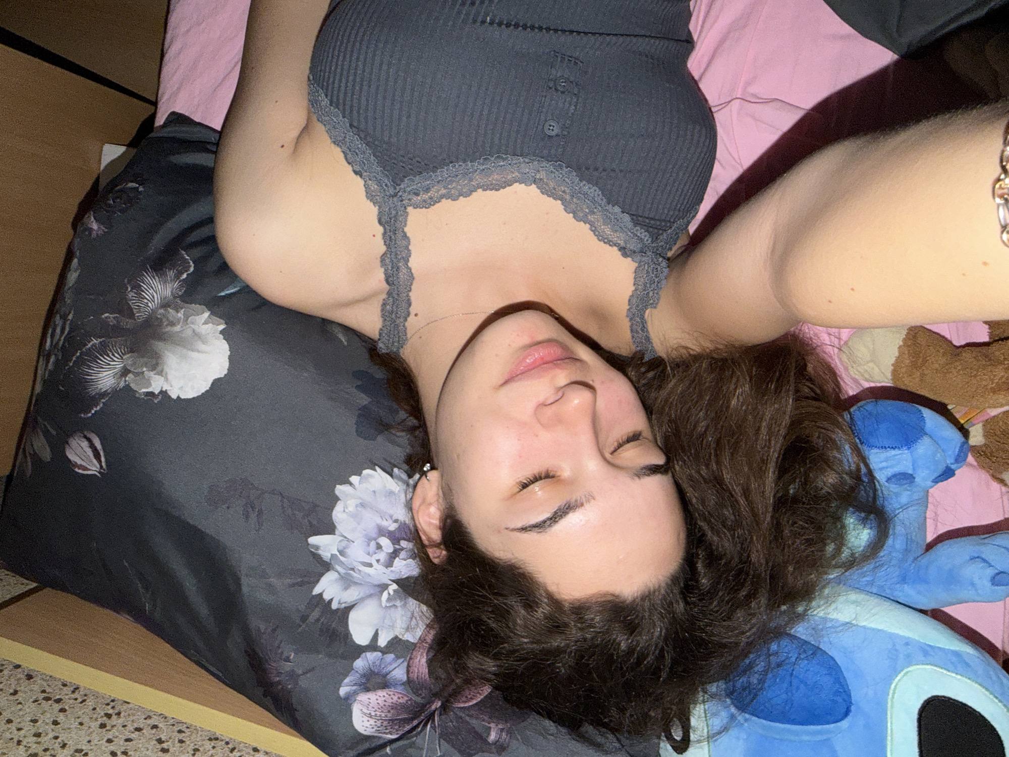The image captures a young woman lying on her back, appearing to be resting or sleeping, in what looks like a bedroom in Sofia, Bulgaria. Her head is tilted to the left side of the frame, with her dark, curly hair fanned out on a pillow. Her eyes are closed, and her lips are slightly parted, conveying a sense of peace or relaxation. She has a natural complexion with visible minor blemishes and defined eyebrows.
She is wearing a dark grey, ribbed tank top with delicate lace trim along the neckline and straps. A thin silver chain necklace is visible on her chest, and a silver-colored chain bracelet is on her right wrist, which is extended upwards out of frame. An earring can be seen in her left ear.
She is lying on a bed with pink sheets. A dark grey pillow with prominent white and purple floral patterns (resembling peonies and irises) supports her head. To her right, nestled beside her head, is a blue stuffed animal resembling the character Stitch from Disney's Lilo & Stitch, along with a glimpse of a brown stuffed animal, possibly a teddy bear. The lighting suggests an indoor setting, likely evening or night, with artificial light casting some soft shadows. A wooden floor or bedside table is visible on the left side of the frame. There is no visible text in the image.