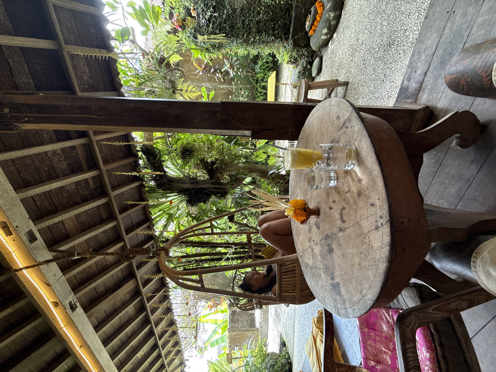 A medium indoor-outdoor shot looking up and across a circular table and a person in a swing chair in the Ubud District, Indonesia.
A round, wood table sits to the right of the frame. On the table are a glass of yellow liquid, two clear glasses, and a small dark vase with yellow flowers. A person is reclined in a wicker hanging chair behind the table. The chair is hanging from the dark brown exposed beams of the ceiling. The ceiling features lights along a beam. The background is a lush green outdoor courtyard, with plants and a stone bench. A wooden porch is visible to the right and left.