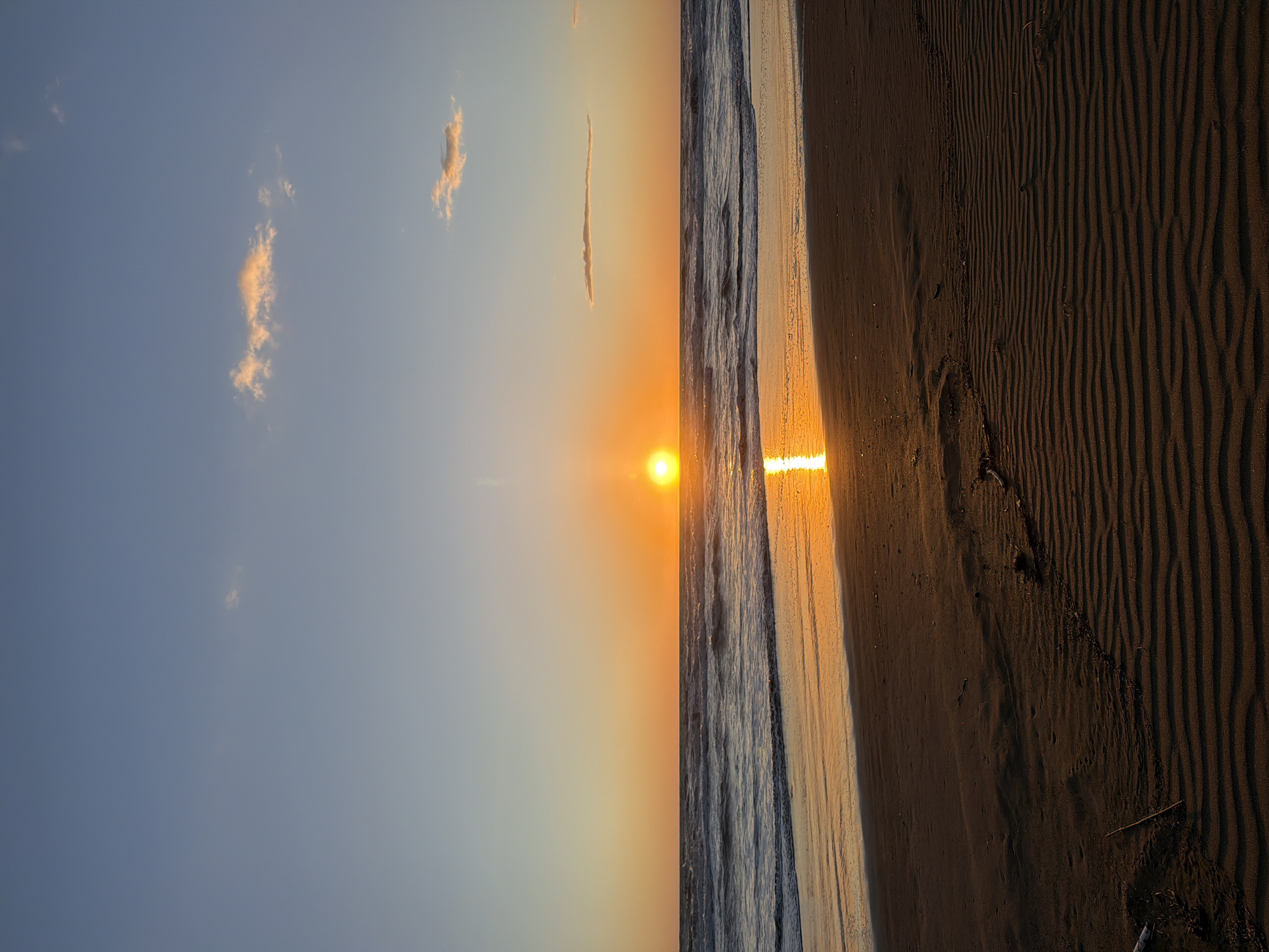 The image depicts a sunset at a beach in Castagneto Carducci, Italy. The sun is positioned on the horizon, reflecting on the water. To the right of the water is a sandy beach, textured with wave-like patterns. To the left of the water is the sky with a few clouds. The image appears to be rotated 90 degrees counterclockwise. There are no people visible in the scene.