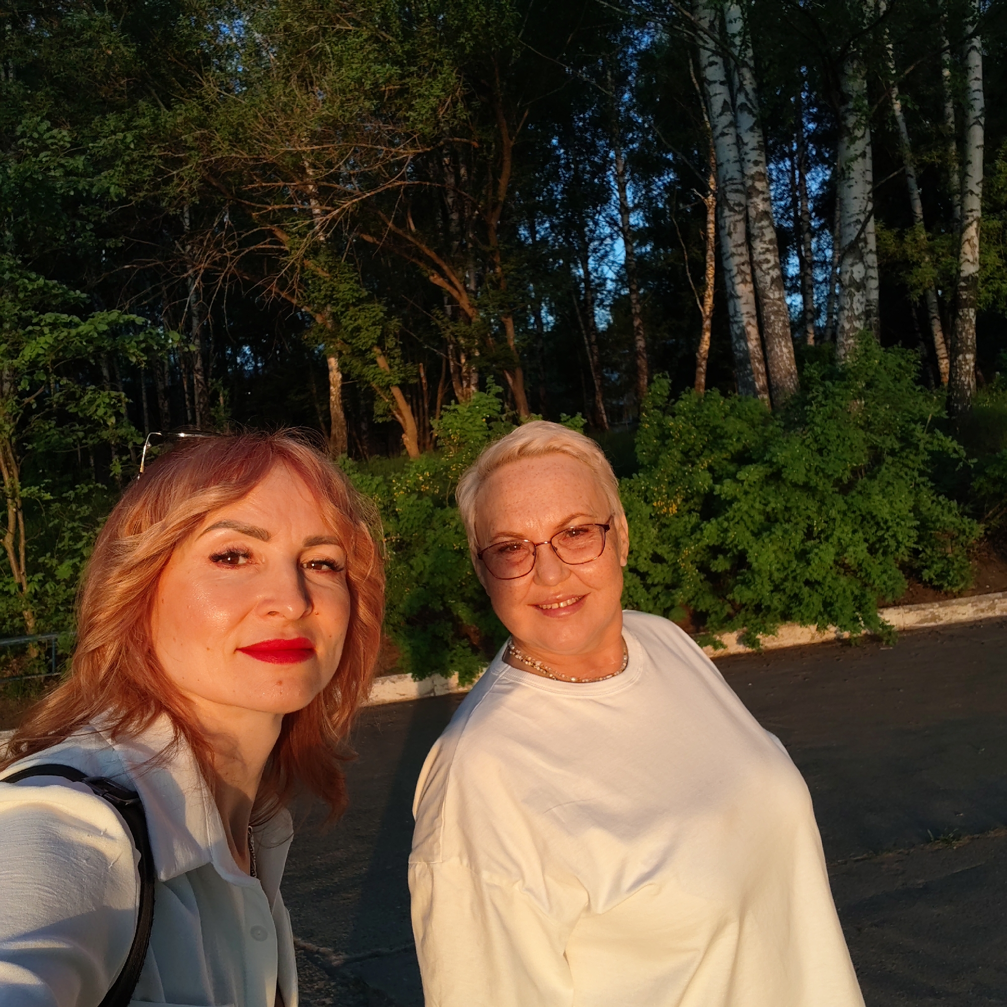 Two women are posing for a picture in front of a wooded area. The woman on the left has red hair and is wearing a white shirt. The woman on the right has short, blonde hair and is wearing glasses and a white t-shirt with a beaded necklace. Behind them is a row of trees, some with white bark. The location is Chaikovsky, Russia.
