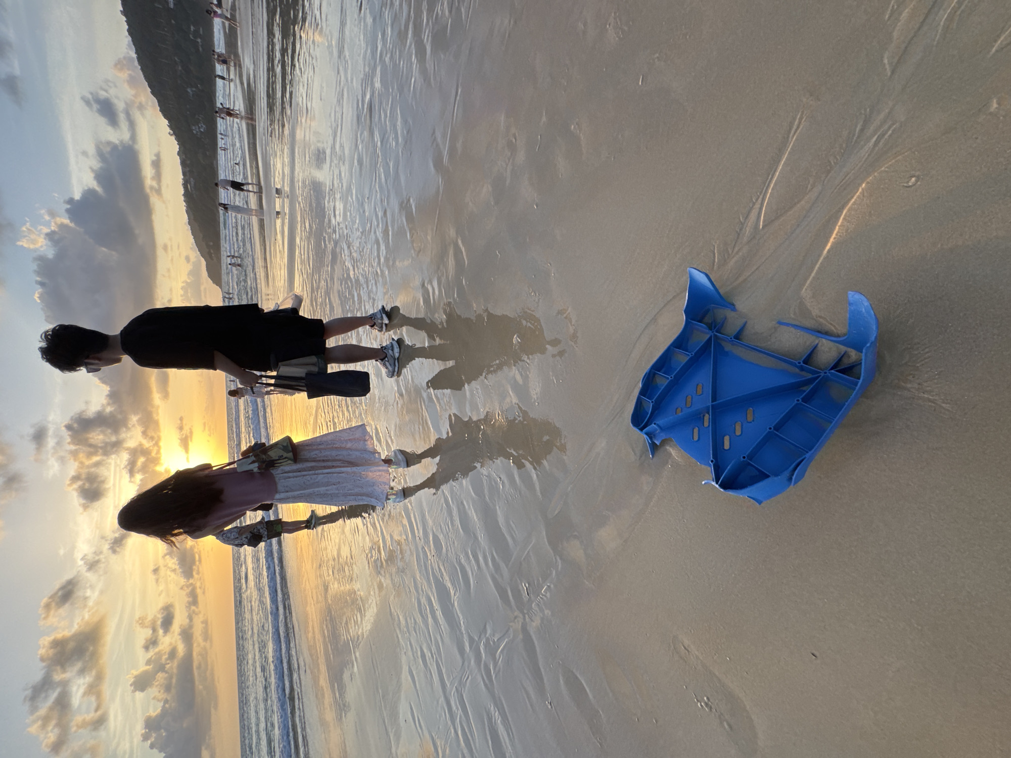 At dusk on Karon Beach in Karon, Thailand, a blue plastic object, possibly a discarded toy or piece of equipment, rests on the wet sand in the foreground. Two individuals, a woman with long dark hair wearing a pink top and a long white skirt, and a man in a black t-shirt and dark shorts, stand near the water's edge in the middle ground, facing the ocean. Their reflections are visible in the shallow water covering the sand. In the background, other people are scattered along the beach and in the ocean, enjoying the sunset. The sky is a gradient of orange and yellow, with scattered clouds, indicating the sun is setting behind the hills that line the coast. The waves gently lap the shore, leaving ripples and patterns in the wet sand.