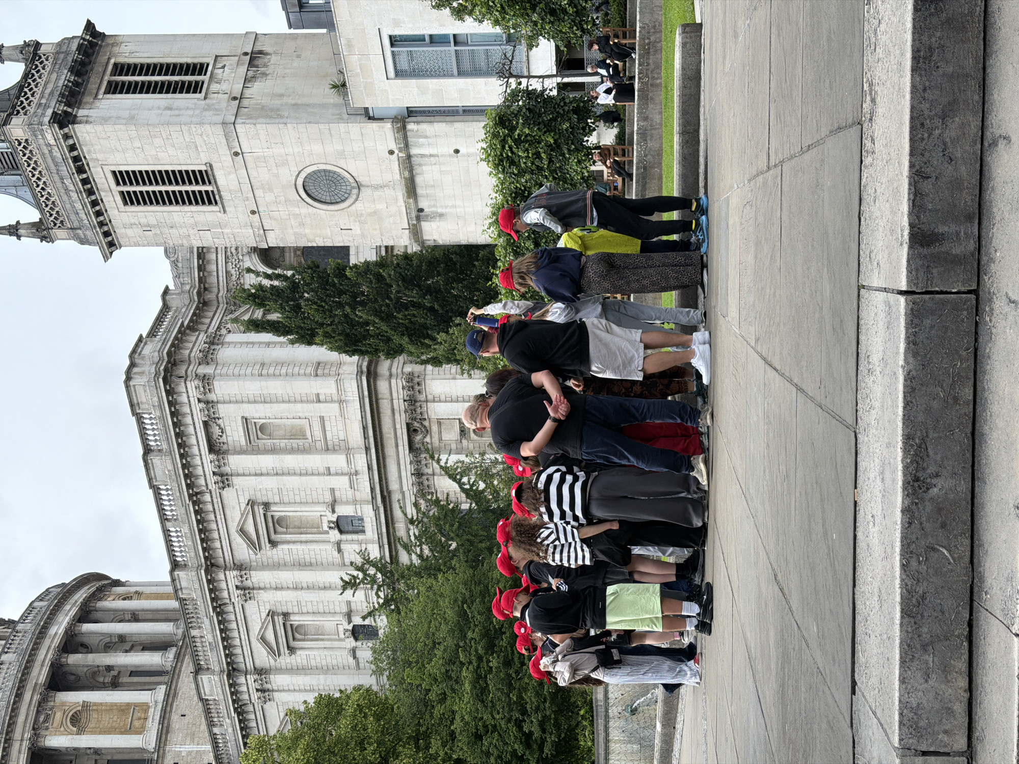 The image depicts an outdoor scene in the city of London, United Kingdom, featuring St. Paul's Cathedral and surrounding architecture. In the foreground and midground, a group of approximately 15-20 individuals is gathered on a paved stone area. Many of the younger individuals in the group are wearing red baseball caps and casual attire such as t-shirts, shorts, and hoodies. An adult male in a black t-shirt and dark pants stands with his back to the viewer, appearing to address or interact with the group. Another adult male in a black t-shirt and white shorts stands to the right, holding a blue bottle and a phone, seemingly taking a picture or video. Several other adults are interspersed within the group.
The background is dominated by architectural structures. St. Paul's Cathedral's classical stone facade, featuring columns and a domed structure, occupies the upper left portion of the image. To its right, a prominent stone clock tower or steeple with louvered vents and a round window is visible. Further to the right, a modern stone building with large windows is present. Between these structures and the group of people, there are several large green trees and a manicured grassy area with stone benches where a few people are seated. The sky is overcast. The ground in the immediate foreground consists of rough-hewn stone steps leading up to the large, grey flagstone pavement where the group stands. A water feature is partially visible on the far left.