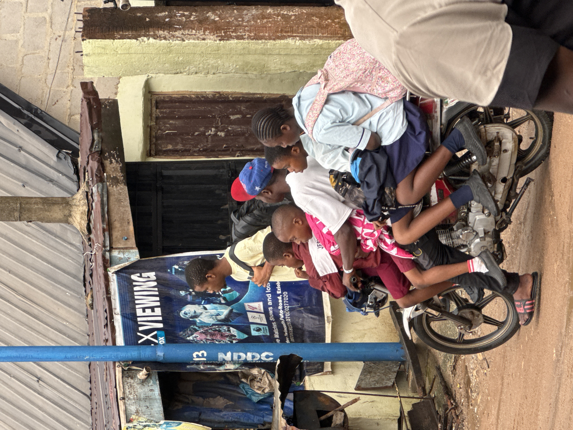 In Ihiagwa, Nigeria, several children are crammed onto a motorcycle parked along a weathered street. The children wear various casual and school-style clothes. A blue utility pipe, marked "13 NDDC", runs horizontally across the lower left of the frame, in front of a weathered wall and a sign advertising "X Viewing" for live matches and shows. The street appears gritty and somewhat unkempt, with rusted metal elements visible.