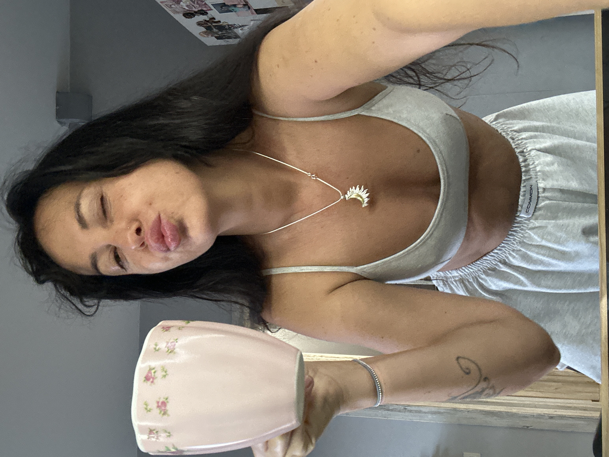 A young woman with long dark hair is taking a selfie while holding a pink mug decorated with small pink flowers. She is wearing a grey bralette and grey shorts. She is winking and puckering her lips for the photo. A silver necklace with a sun pendant is visible around her neck. She is also wearing a silver bracelet on her left wrist. A tattoo is visible on her left forearm. The background appears to be an indoor setting, possibly a bedroom or living area, with a mirror in the foreground reflecting part of the scene. There are some pictures or postcards on the wall in the background. The scene does not contain any specific landmarks or indicators that would place it in Irkutsk, Russia.
