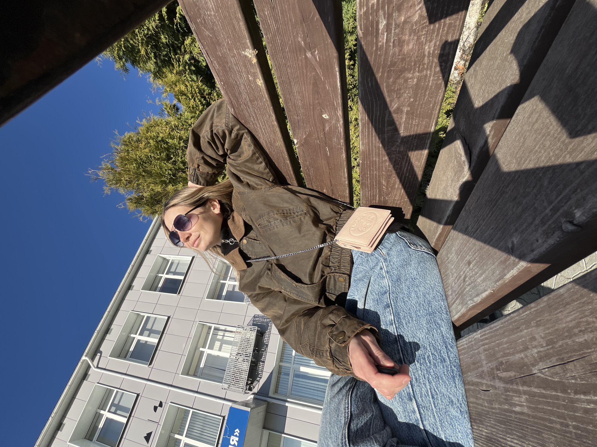 A woman wearing sunglasses, a brown jacket, and jeans is sitting on a wooden bench. She has a small pink purse slung over her shoulder. Behind her is a light gray building with multiple windows. A tree is visible in the background, and the sky is clear and blue. Based on the Cyrillic script visible on a sign, the image was taken in the city of Chaikovsky, Russia.