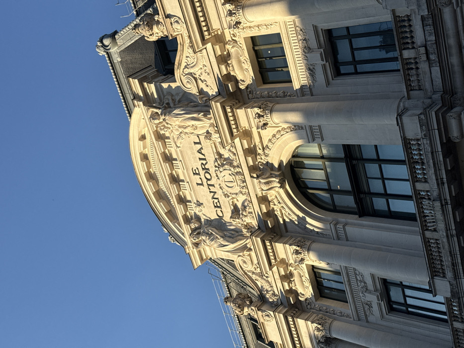 The image displays an upward view of the ornate limestone facade of the Le Centorial building, situated at 33 Rue de Miromesnil, 75008 Paris, France. The building exhibits a Neoclassical architectural style with extensive decorative elements. Centrally located on the upper visible floor is the inscription "LE CENTORIAL" in capital letters. This inscription is flanked by two large, draped allegorical female sculptures standing on pedestals, with additional smaller bas-reliefs below them. The facade features fluted columns with ornate Corinthian capitals, supporting an elaborate entablature and cornice work. Multiple large windows are visible, including a prominent arched window and several rectangular multi-paned windows. Reflections are present in the window panes, and interior venetian blinds are partially visible through some. A stone balustrade with multiple balusters runs across a lower section of the building. Portions of a dark mansard roof with decorative cresting are visible at the top of the structure. Strong, directional sunlight illuminates the building from the left, casting deep shadows that emphasize the architectural contours and sculptural details. The sky above is clear and uniformly blue. No individuals, actions, or specific interactions are depicted.