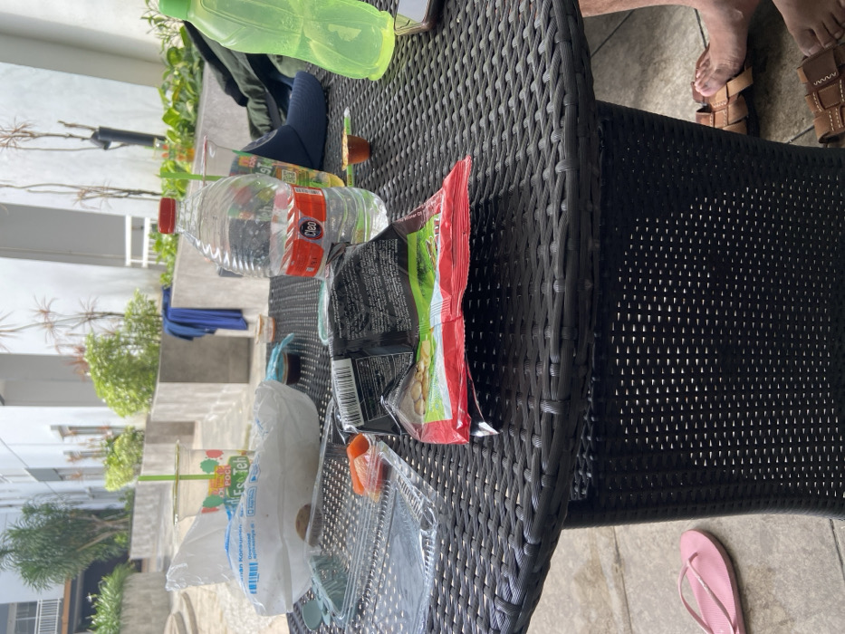 A rattan table is littered with personal belongings. A red and white water bottle, a green bottle, a bag of chips, a cup, a takeout container, and a blue sock are on the table. In the background, there is some greenery, a building, and stairs. A pair of sandals and bare feet are visible on the upper right side of the image. A pink flip-flop is on the floor next to the table.