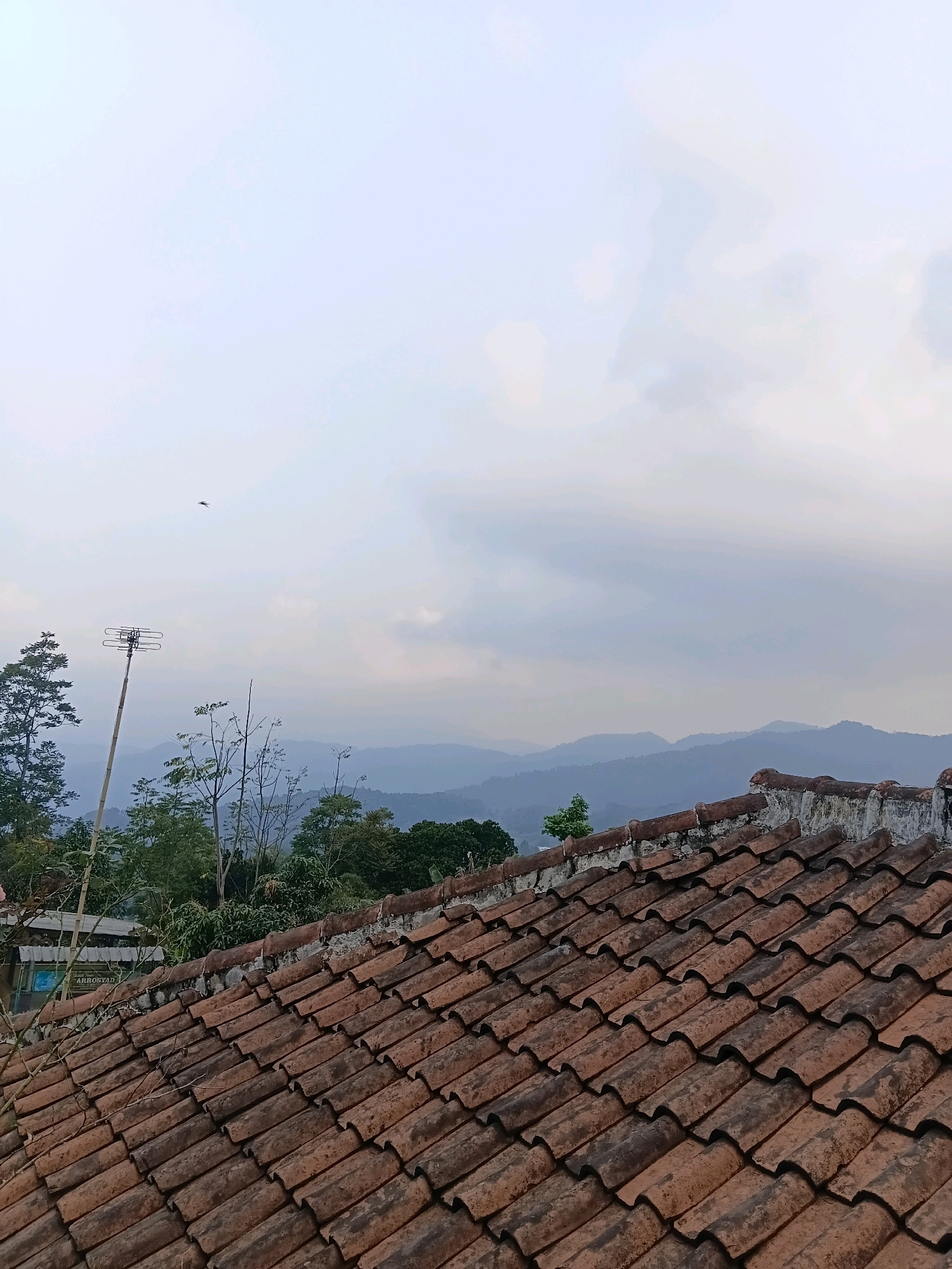 The image depicts a rooftop view in Ciater, Indonesia, overlooking rolling hills under a cloudy sky. In the foreground, a weathered terracotta tiled roof occupies the lower half of the frame, with its uneven surface suggesting age and exposure to the elements. To the left, a tall bamboo pole supports an old television antenna. Beyond the rooftop, lush green trees and foliage form a dense line, partially obscuring buildings with signage, including one that reads "ARROSYAD." The background reveals a series of receding, hazy blue hills, characteristic of a mountainous landscape. The sky is overcast with diffused light, hinting at either dawn, dusk, or a heavily clouded day. A small, dark object, possibly a bird or insect, is visible in the upper left portion of the sky.