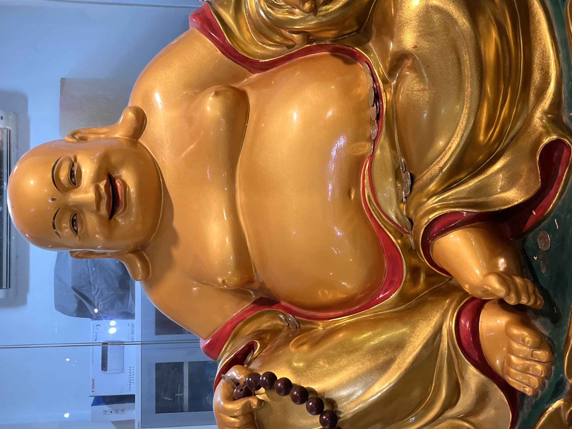 A golden statue of a laughing Buddha is the central focus of this image. The Buddha is depicted with a round, smiling face, large ears, and a prominent belly, characteristic of the Hotei figure in Buddhist tradition. He is seated, draped in flowing golden robes with red trim, and is holding a string of dark brown prayer beads in his left hand. Several coins are scattered around his lap and the base of the statue. The background is blurred, but appears to be an indoor setting with shelves and what looks like a printer box on one of them. The lighting is bright, highlighting the metallic sheen of the statue. The image appears to be taken at a medium close-up, emphasizing the details of the statue. Without additional information, the specific location within Colombo, Sri Lanka cannot be determined from the image alone.