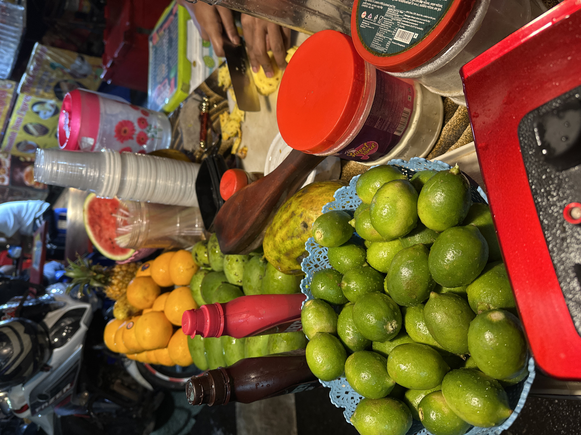 The scene depicts a street food stall in Dhaka, Bangladesh, captured at night under artificial illumination. The primary focus is on a display of fresh fruits and preparation activity.
In the immediate foreground, a blue plastic basket holds a quantity of wet, green limes. To its right, a partial view of a red surface, possibly a counter or machine, is visible. Behind the limes, two upright squeeze bottles are present: one dark brown and one red. A large mound of additional green, round fruits is situated behind these bottles.
Central to the image is a vibrant array of fruits: a substantial pile of bright orange citrus fruits (likely oranges or mandarins), a whole pineapple, and a large slice of watermelon with its red flesh exposed.
To the right, a stack of clear plastic cups is visible, alongside a pink and white floral-patterned drinking bottle. A wooden pestle or similar tool rests on the surface. Multiple plastic containers with red lids are present, with one container clearly labeled "Sweet Land."
In the upper right, a pair of human hands is actively engaged in slicing a yellow fruit, identified as a pineapple, on a cutting surface with a silver knife. Smaller, already cut pieces of pineapple are scattered nearby.
The background on the left reveals the front section of a parked motorcycle with a dark helmet placed on its seat. The overall environment suggests an outdoor market or street vendor setup, characterized by various objects and equipment common to such operations, illuminated by overhead lights. The ground or immediate surface appears dark.