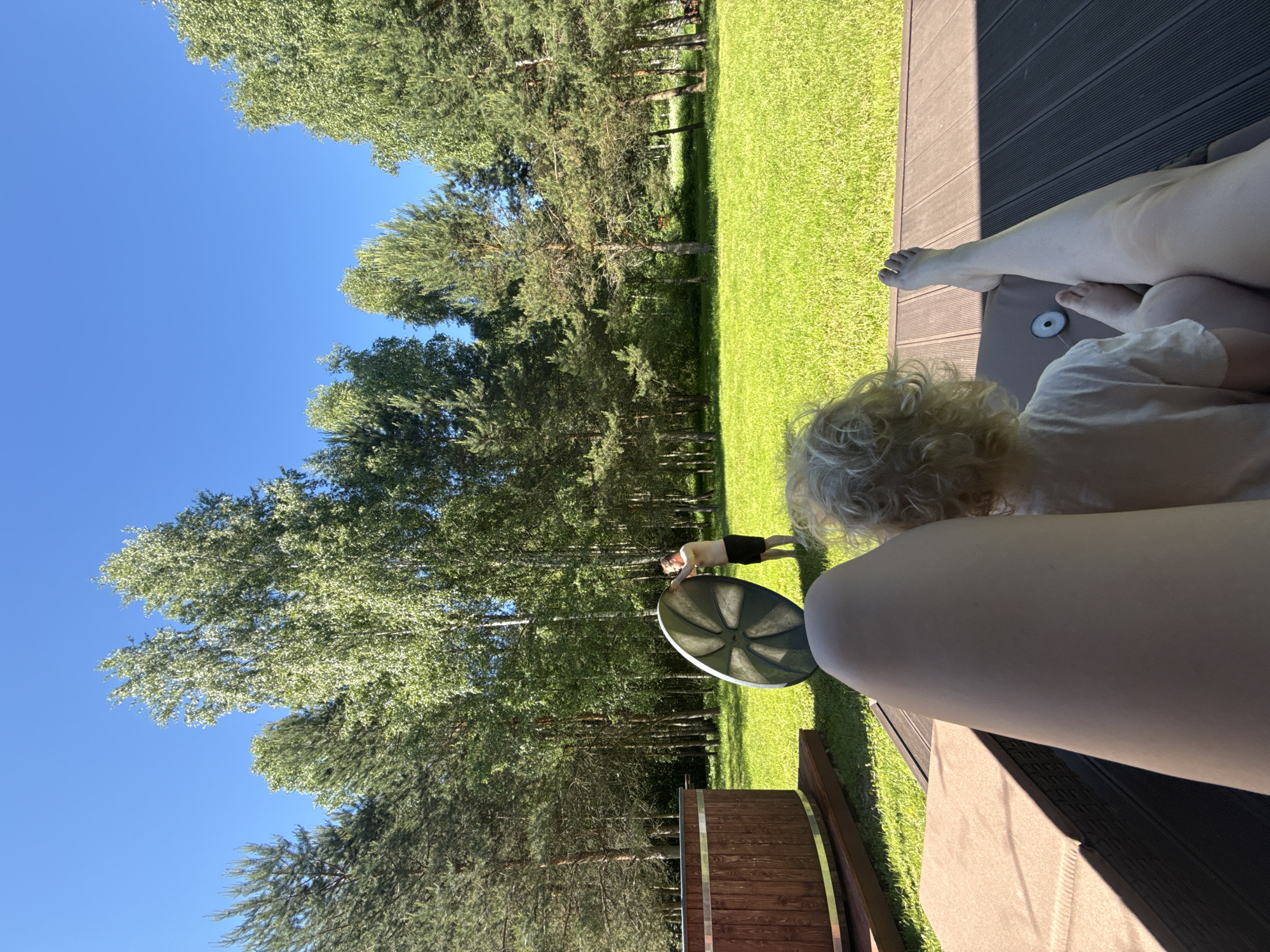 An outdoor, eye-level shot captures a bright, sunny day in what appears to be a rural or forested residential area, possibly in Копнино, Russia. The image is taken from the perspective of someone seated, with their legs and bare feet visible in the lower right foreground, resting on a dark wooden deck or patio. To the left of these legs, a small child with curly blonde hair is visible from behind, seemingly looking out towards the scene.
The immediate midground features a wooden hot tub with metal banding, and a large, dark, possibly metallic, round object with a segmented pattern is prominently placed between the hot tub and the child.
In the background, a man, shirtless and wearing dark shorts, is visible standing on a lush green lawn. He appears to be bending over slightly or engaged in some activity. The lawn stretches out towards a dense line of tall trees, including both coniferous and deciduous varieties, forming a natural barrier. The sky above is a clear, bright blue, indicating good weather. The sunlight casts distinct shadows, suggesting it's likely midday or early afternoon. The overall atmosphere is one of relaxation and outdoor activity on a summer day. No visible text is present in the image.