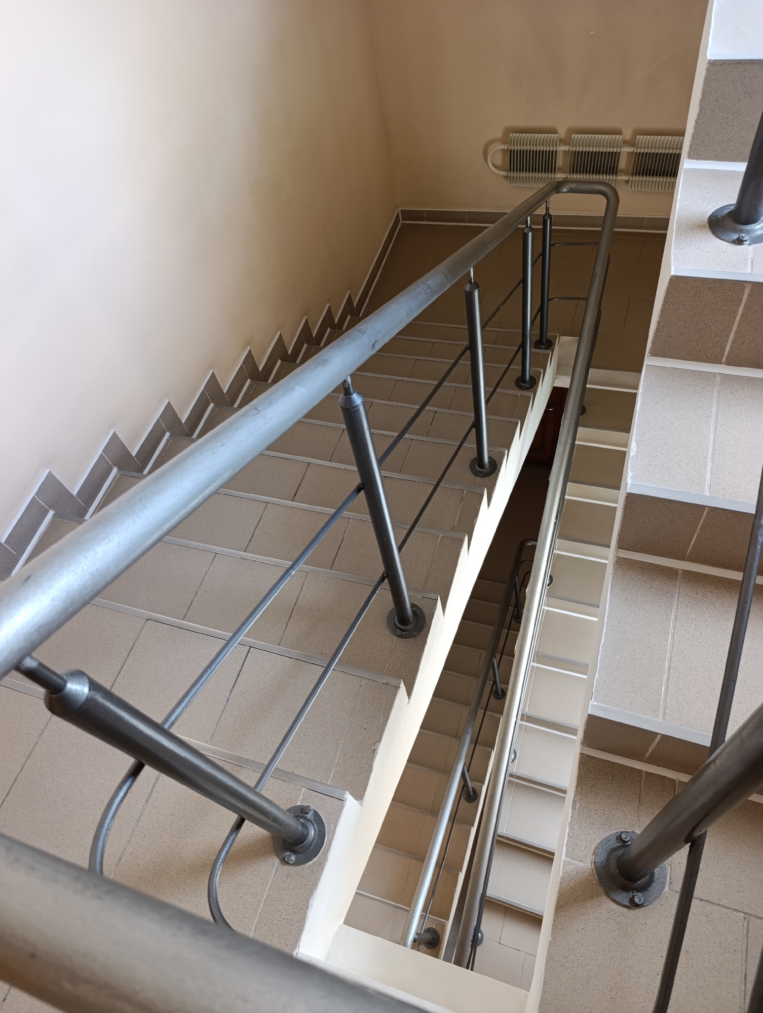 The image shows a view looking down a concrete and tile staircase with metal railings. The walls are painted a light beige color. In the upper right of the image, there is a section of wall with a radiator attached. The staircase appears to be indoors, possibly in a public building or apartment complex. There are no people visible in the image. The lighting suggests it is daytime. The environment is clean and well-maintained. This scene is located in the city of Dubăsari town council, Moldova.