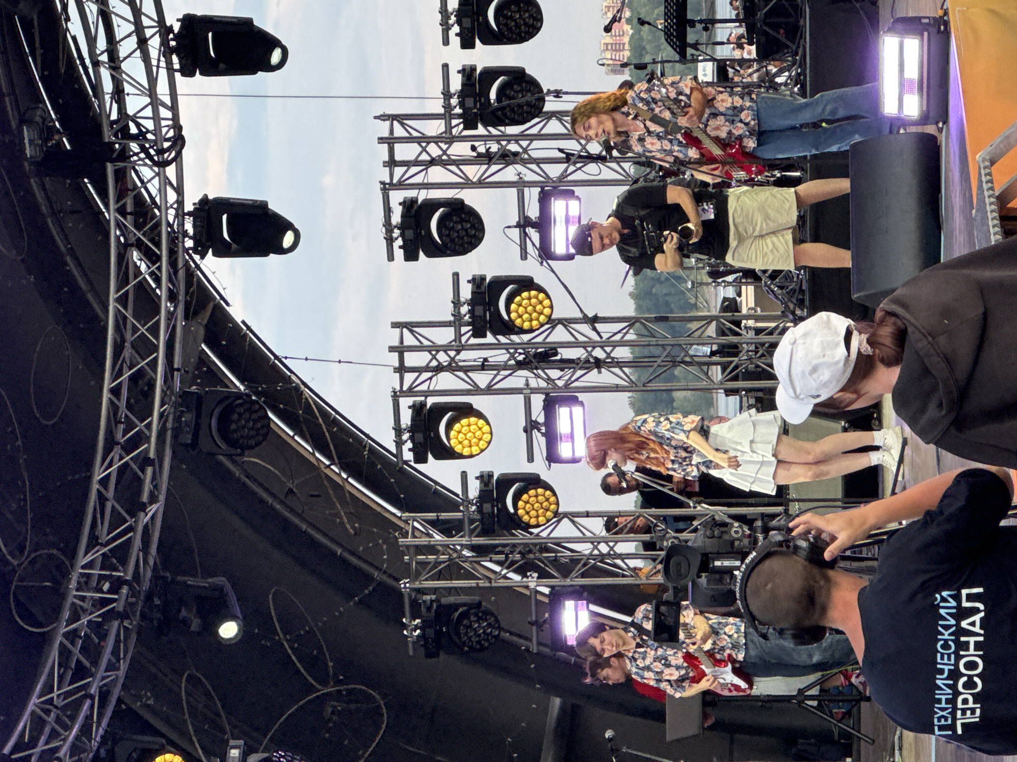 On an outdoor stage in Novosibirsk, Russia, a musical performance is underway. The stage is equipped with multiple lighting rigs and sound equipment, including large speakers. A band, consisting of at least three musicians, is performing. One musician is playing a red electric guitar and singing into a microphone. Another musician, in a floral shirt, is also playing an electric guitar. A third musician, wearing a white skirt and floral top, is singing. Behind the band, a cameraman with a professional video camera is filming the performance. In the foreground, a person wearing headphones and a black t-shirt with the Russian text "ТЕХНИЧЕСКИЙ ПЕРСОНАЛ" (Technical Personnel) is visible, indicating their role in the event. Another person in a white hat is partially visible in the lower right corner. The sky above is overcast, suggesting a cloudy day. The overall scene depicts a live music event, possibly a festival or concert, taking place in an urban outdoor setting.