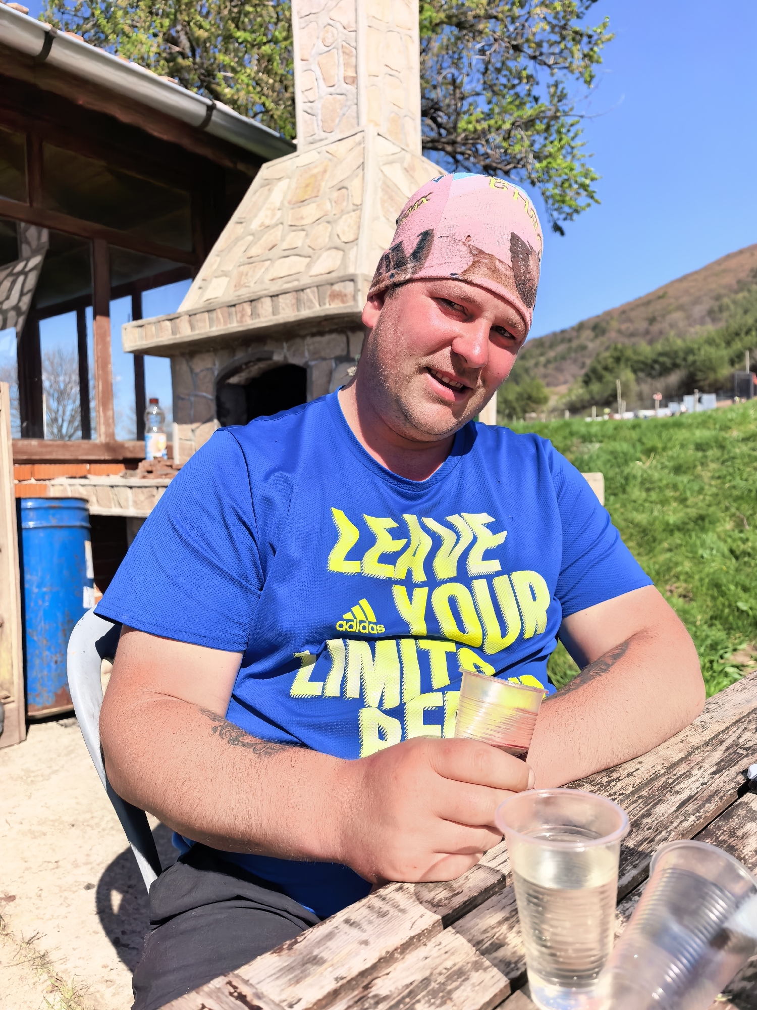 A man is sitting at a wooden picnic table outdoors. He is wearing a blue t-shirt with yellow lettering, a pink bandana, and dark shorts. The man is holding a clear plastic cup in his right hand and has another clear plastic cup on the table in front of him. Behind him is a structure that appears to be an outdoor fireplace or oven constructed of stone. To the left of the man is a blue barrel and part of a building with windows. In the background, there is a grassy hillside and a clear blue sky.