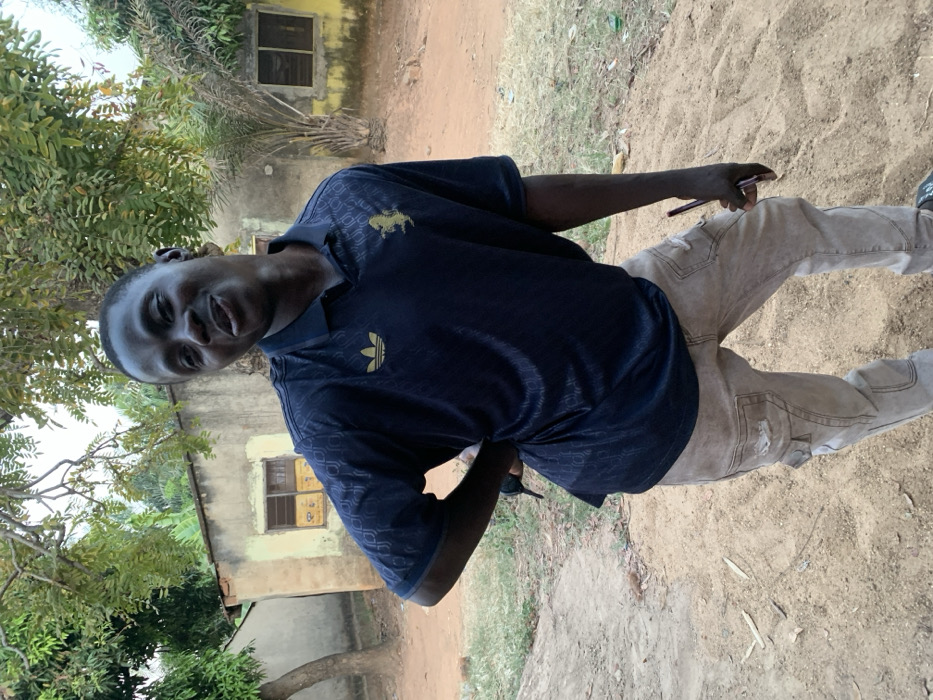 A smiling man stands outside, angled slightly towards the camera. He wears a dark blue polo shirt with gold logos and light-colored jeans with distressed patches. In his hand, he holds a dark-colored object, possibly a phone. The background features a dirt ground, a building with small windows, and trees with green foliage. The image is slightly rotated.