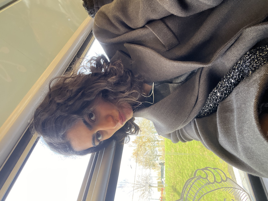 The image depicts a female subject, approximately 20-30 years old, situated within a public transport vehicle, presumed to be in Torcy, France. The subject, with dark, medium-length curly hair, dark eyes, and full eyebrows, is centered and looking directly into the camera with pouted lips. She is wearing a dark grey, possibly wool or wool-blend, overcoat with prominent lapels, under which a lighter grey patterned garment is visible. White wired earbuds are present in her ears, with the wire trailing downwards. A black, textured or sequined, bag strap diagonally crosses her upper torso. The background, viewed through the vehicle's large window, shows an exterior landscape with a bright green grassy embankment, deciduous trees displaying autumnal yellow and orange foliage, and a faint view of a road with a light-colored vehicle in the distance. A white, abstract, wire-frame-like graphic or reflection is partially visible on the lower-left section of the window. The vehicle's interior features a light-colored ceiling and dark window trim.