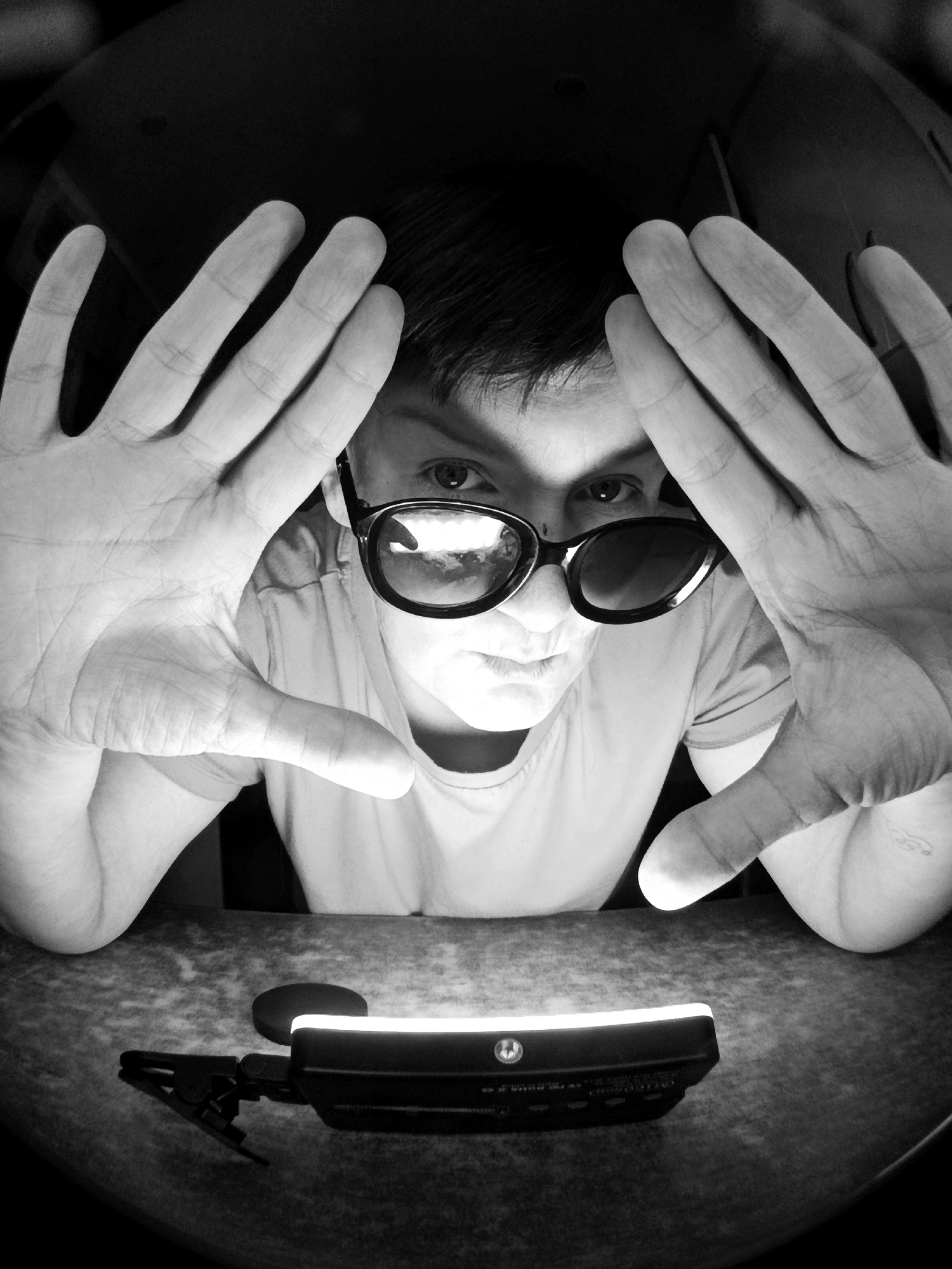 A black and white, high-angle, close-up shot shows a person wearing dark-rimmed glasses, holding their hands up in front of their face, as if to block the camera. The person is looking directly at the camera with a serious expression. Their hands are positioned to obscure most of their face, with only their eyes and mouth visible. In the foreground, a small, black object with a clip is resting on a textured surface. The background is dark and out of focus. The scene appears to be indoors, with no specific location in the city of Фоки, Russia, identifiable.