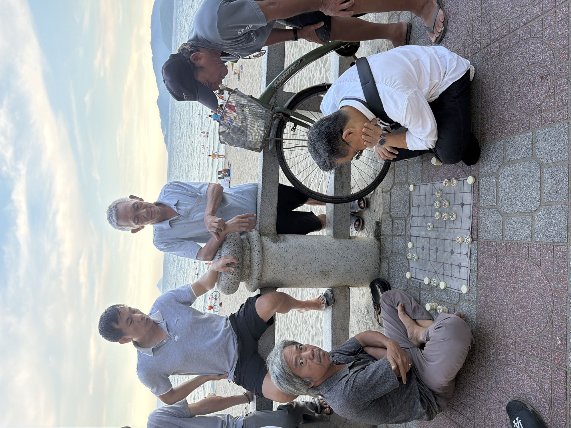 Six men are gathered by the sea in Nha Trang Ward, Vietnam. A game of Xiangqi (Chinese chess) is in progress on a mat on the ground, with pieces set up on the board. One man, wearing a gray shirt and pants, sits on the ground with his arms crossed, looking at the camera. Another man in a white shirt and black pants is crouched down, contemplating the game. Two other men, one in a gray polo shirt and black shorts and another in a light blue polo shirt, are standing behind a stone railing. A man in a dark gray t-shirt and shorts is leaning over the railing, looking at the chessboard. A bicycle with a basket is parked next to the railing. The background shows a beach with people swimming and relaxing, and the sea with hills in the distance. The sky is partly cloudy. The ground is paved with interlocking square tiles in shades of gray and red.