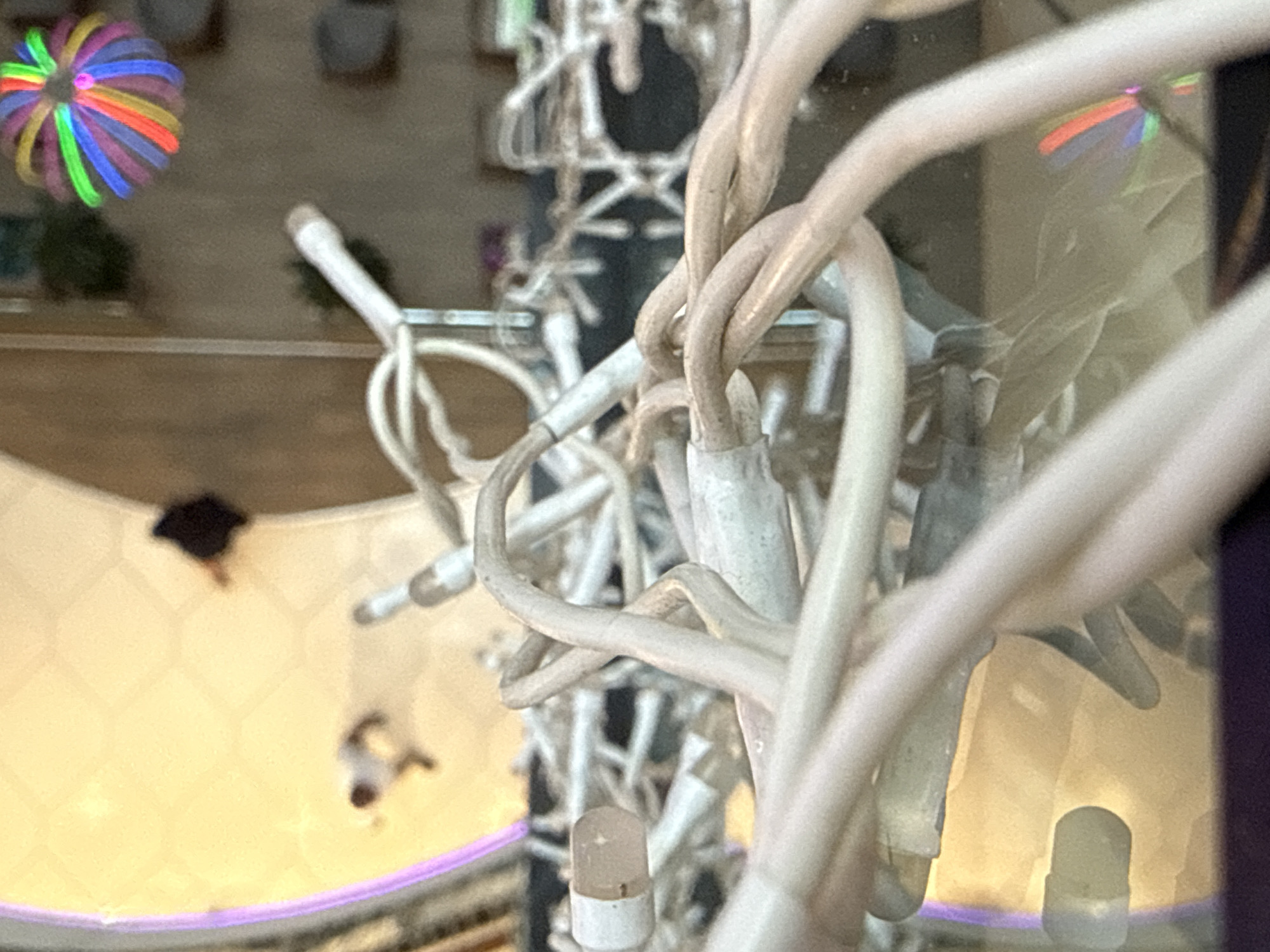The image shows a close-up view of tangled white Christmas lights, obscuring much of the background. In the blurred background, several figures can be faintly seen. One figure, dressed in dark clothing, appears to be walking. Another figure, dressed in lighter clothing, is also visible. In the upper right quadrant of the image, a colorful, blurry object resembling a multi-colored sphere or pom-pom is suspended in the air. The lighting suggests an indoor setting with artificial lights, and a reflective surface, possibly a floor or table, is visible beneath the lights. The environment appears to be a public space, possibly a shopping mall or a decorated venue. No specific landmarks of Baku, Azerbaijan, are identifiable in the image.