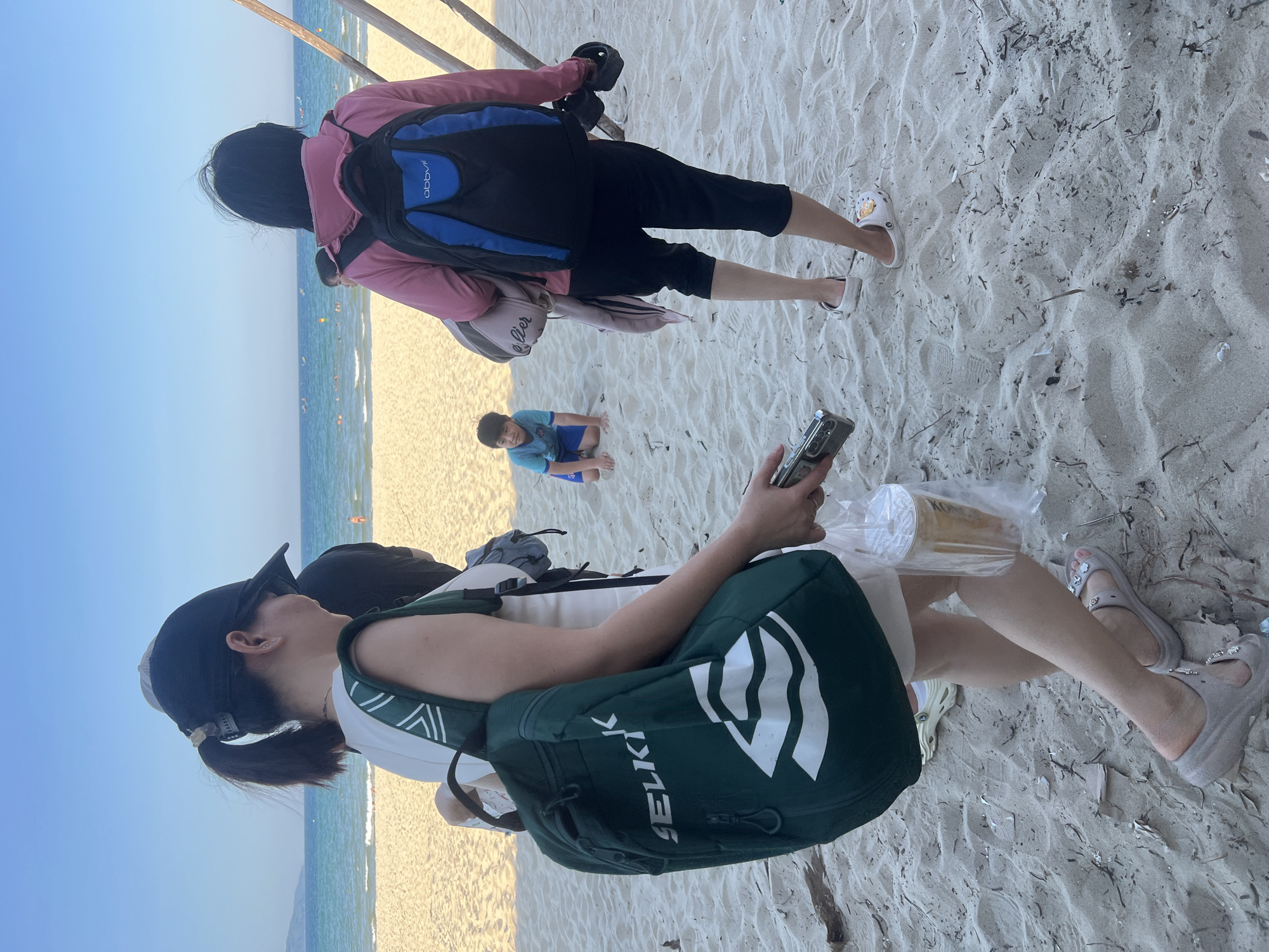 The image shows a group of people on a sandy beach in Da Nang, Vietnam, with the ocean and sky in the background. In the foreground, a woman wearing a black baseball cap and a white top is carrying a green backpack. She is holding a plastic bag with a drink. To her right, another person wearing a pink jacket and a blue and black backpack stands facing away from the camera. Further back on the beach, a young boy in a blue shirt and shorts is kneeling in the sand. The scene is illuminated by natural daylight, suggesting it is daytime.