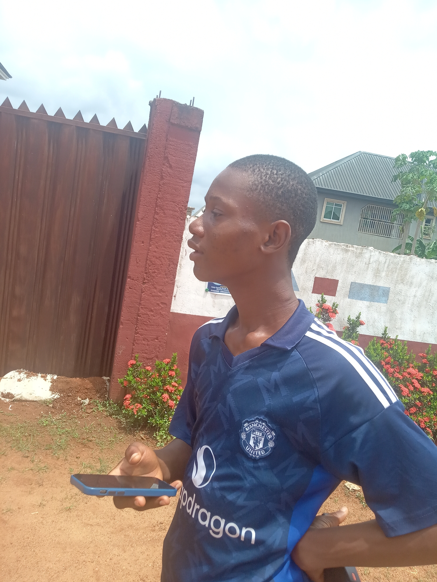 A young man stands outdoors in Ukwu-Abwa, Nigeria, looking towards the left with a neutral expression. He is wearing a dark blue Manchester United football jersey featuring white stripes on the shoulders, the club's crest on the chest, and a partially visible "Snapdragon" logo. In his left hand, he holds a blue smartphone.
The background consists of a tall reddish-brown metal gate topped with pointed spikes. Behind the man, there is a white wall adorned with red and blue rectangular painted sections, partially covered by bushes with small red flowers. A white sign on the wall is partially visible, showing text like "Holy Family Abwa..." and "PHONE: 090...". Further back, a house with a gray corrugated metal roof and white-framed windows is visible. The foreground shows reddish-brown dirt ground. The sky is bright and appears overcast, indicating daytime.