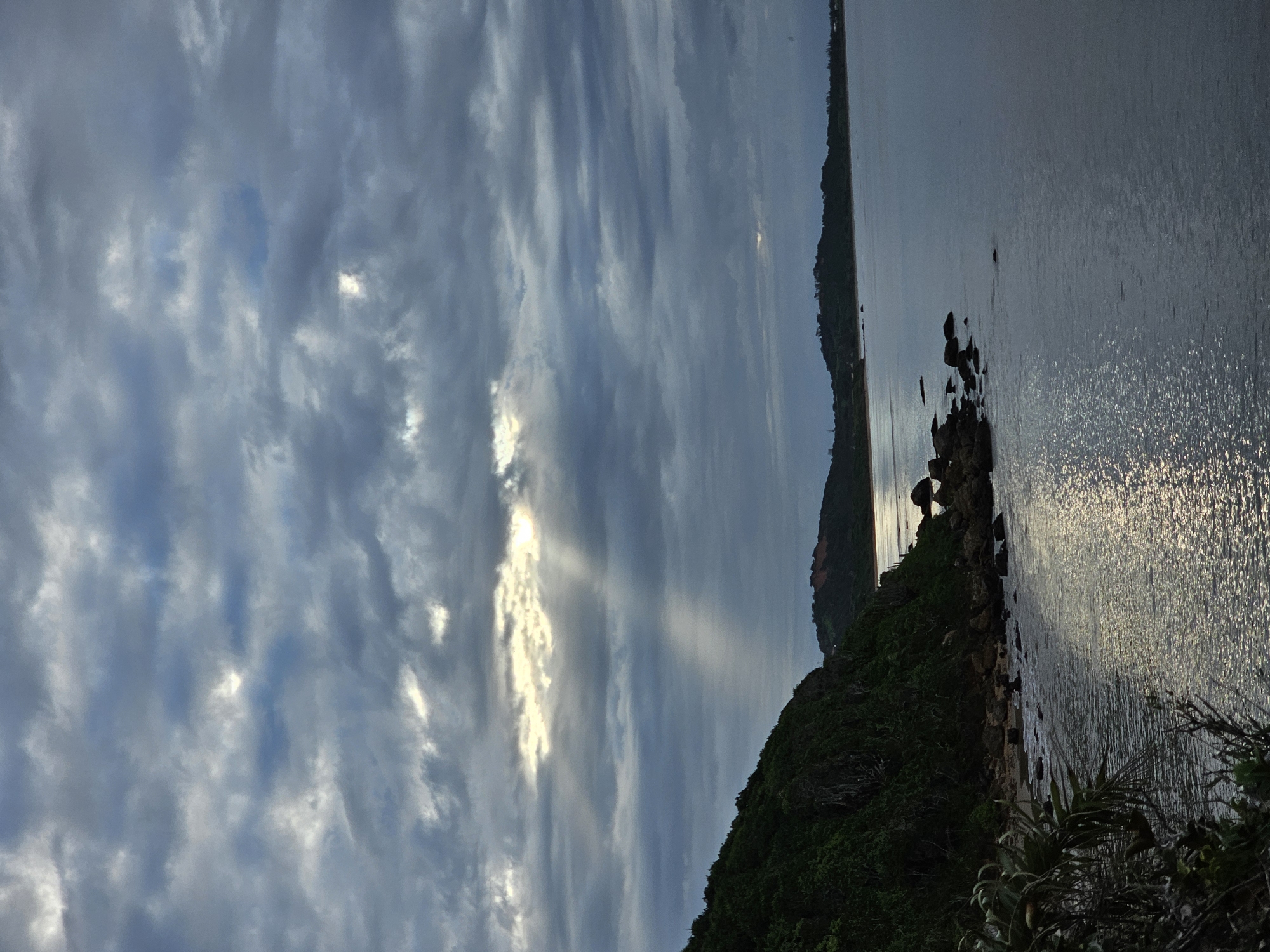 The image depicts a coastline scene in Armação dos Búzios, Brazil. The sky is filled with textured clouds, with a visible sunbeam. The coastline is covered in vegetation and rocks leading to the ocean. The ocean reflects sunlight, creating a shimmering effect on the water's surface.