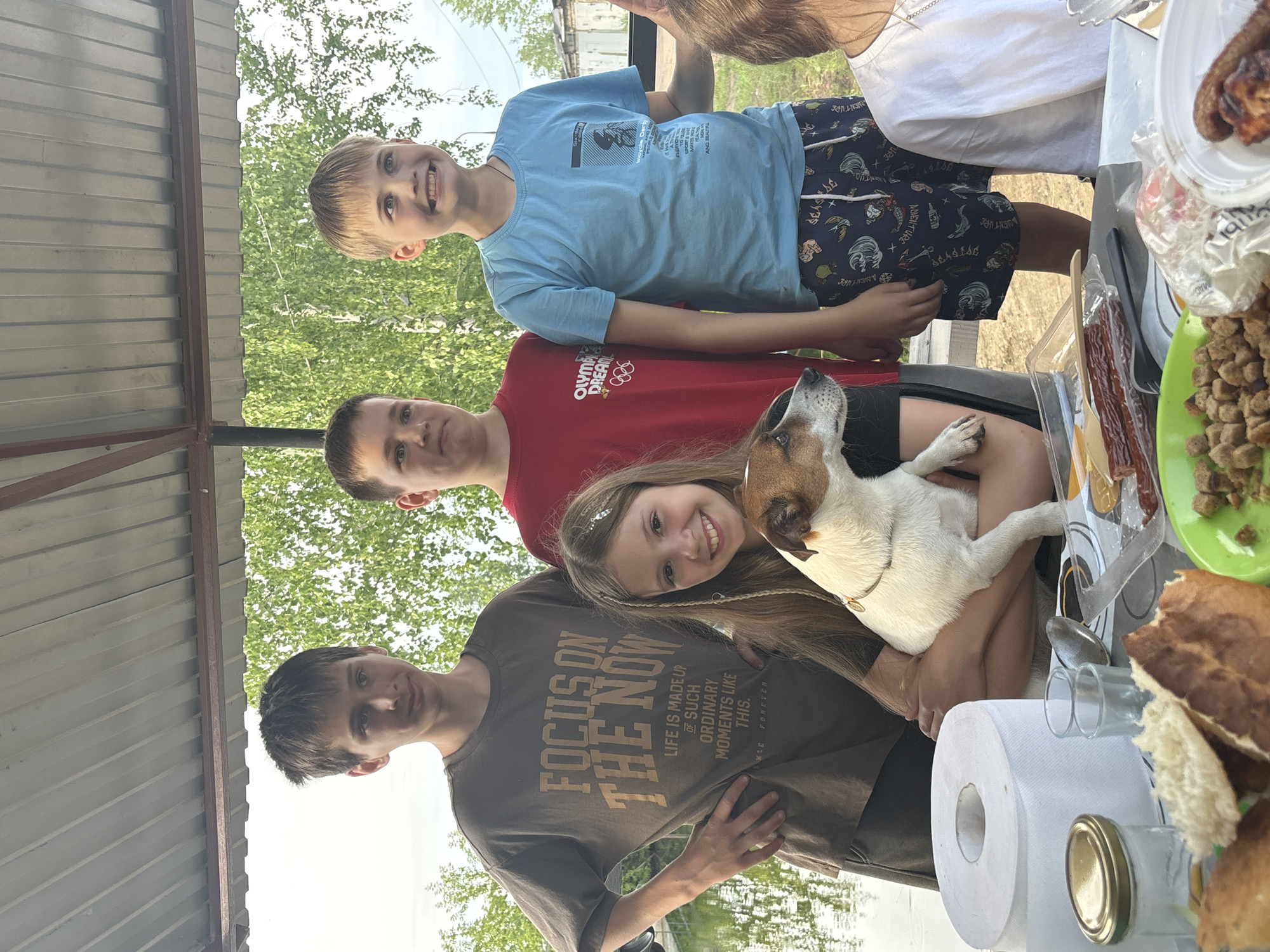 Four individuals, three males and one female, and a dog are gathered outdoors under a covered structure. The male on the far left, appearing to be a teenager, wears a dark t-shirt with the text "FOCUS ON THE NOW". Next to him is another male, also appearing to be a teenager, in a red t-shirt. To his right stands a younger male in a blue t-shirt and patterned shorts. The female, positioned in front of the dog, is holding a Jack Russell Terrier. She has long blonde hair and is smiling. The dog is looking to the right. A table in the foreground is laden with food items including bread, sausages, and what appears to be croutons or small pieces of bread. A roll of paper towels, glasses, and cutlery are also visible on the table. The background shows greenery and trees, suggesting an outdoor setting, possibly near water given the reflection visible. The environment suggests a picnic or gathering in a park or backyard area in Chaikovsky, Russia. The overall atmosphere is casual and familial.