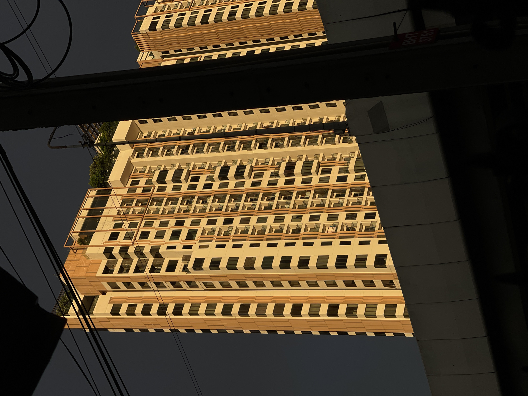 This media file captures a low-angle, upward view of a modern, cream-colored building with numerous windows, set against a dark, overcast sky. The building's facade features repeating horizontal bands of light brown and cream, creating a rhythmic pattern. The windows are mostly dark, with some reflecting the dim light. The building appears to be very tall, with multiple stories visible.
The perspective is unusual, as the building is viewed from below and is tilted. A thick, dark beam runs horizontally across the upper portion of the image, obscuring part of the building and casting a shadow. Below this beam, the building's reflection is visible, mirroring the structure above, suggesting the image might have been taken in relation to a reflective surface like water or glass, or it could be an optical illusion due to the angle.
Several dark, thin wires are strung diagonally across the frame, adding to the urban feel. A small red sign with white text is visible on the underside of the horizontal beam. The text on the sign is partially obscured but appears to read "BHT 3761".
The lighting suggests it is either early morning or late evening, with the sun low in the sky, casting a warm glow on the building. The dark sky and absence of bright sunlight imply potentially cloudy or stormy weather. The presence of power lines and the architectural style of the building are indicative of an urban environment, possibly in Bangkok, Thailand, as suggested by the location context. There are no people or clear activities depicted; the focus is on the architecture and the abstract interplay of light, shadow, and reflection.